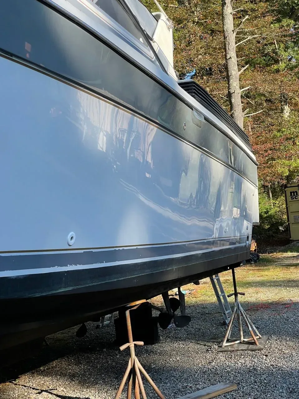 A low-angle view of a white motorboat hull supported by metal jack stands on a gravel lot with trees in the background.