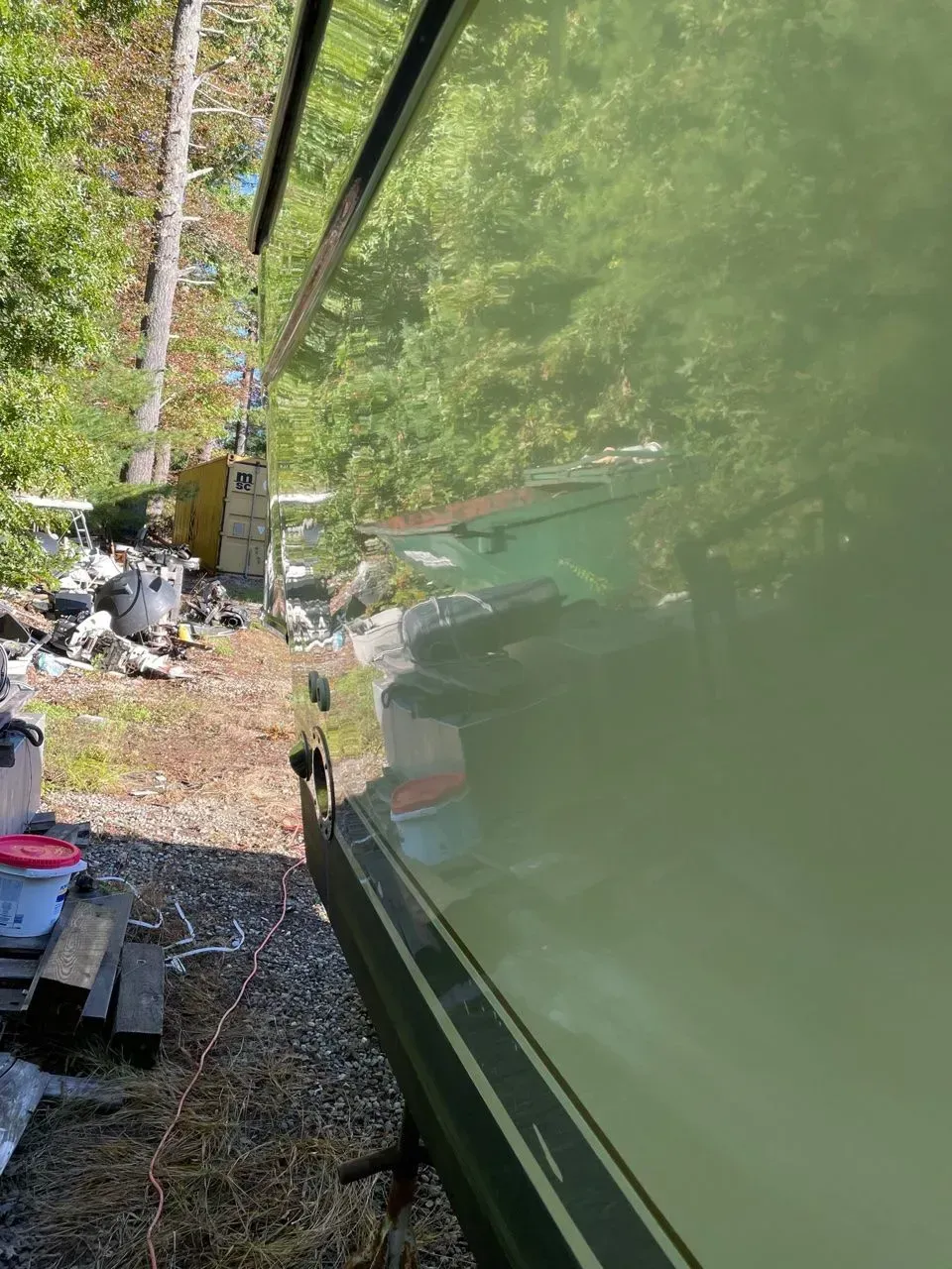 A side view of a glossy, light green boat hull reflecting the surrounding trees and wooded area.