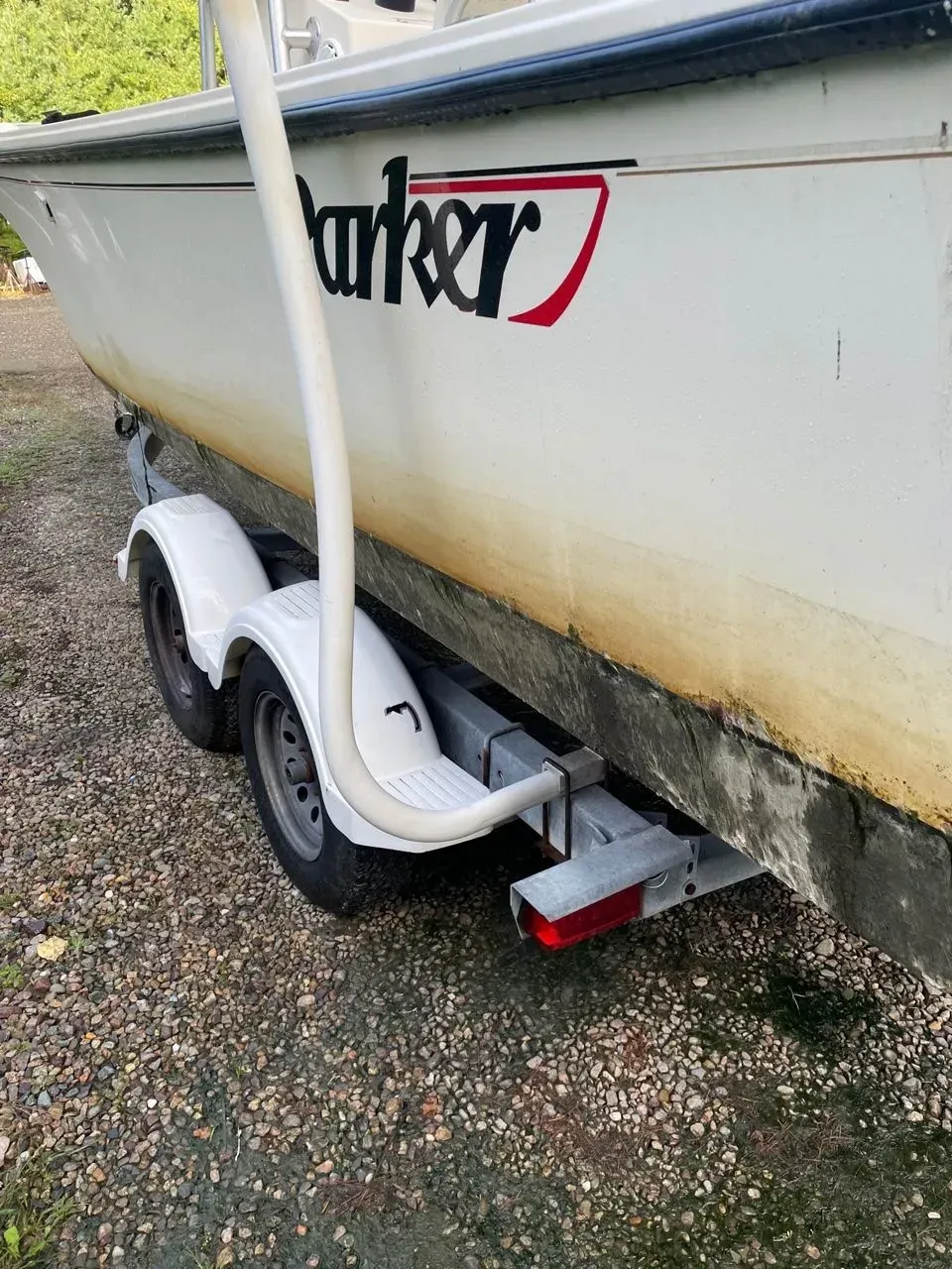 Side view of a white Parker boat on a dual-axle trailer, showing stained hull and a white guide pole on a gravel surface.