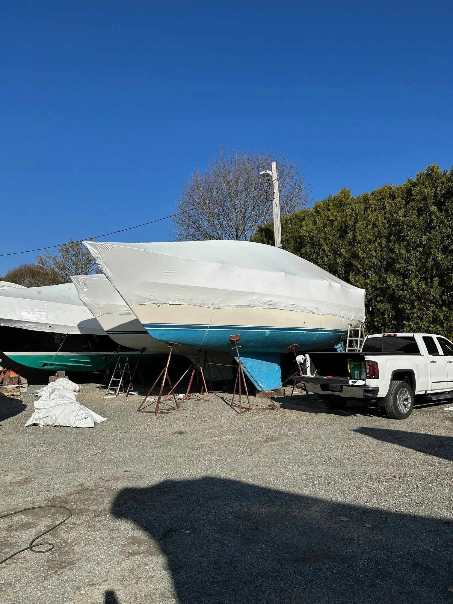 Sailboat on blocks, wrapped in white. Blue hull, gray ground, and white truck in a lot.