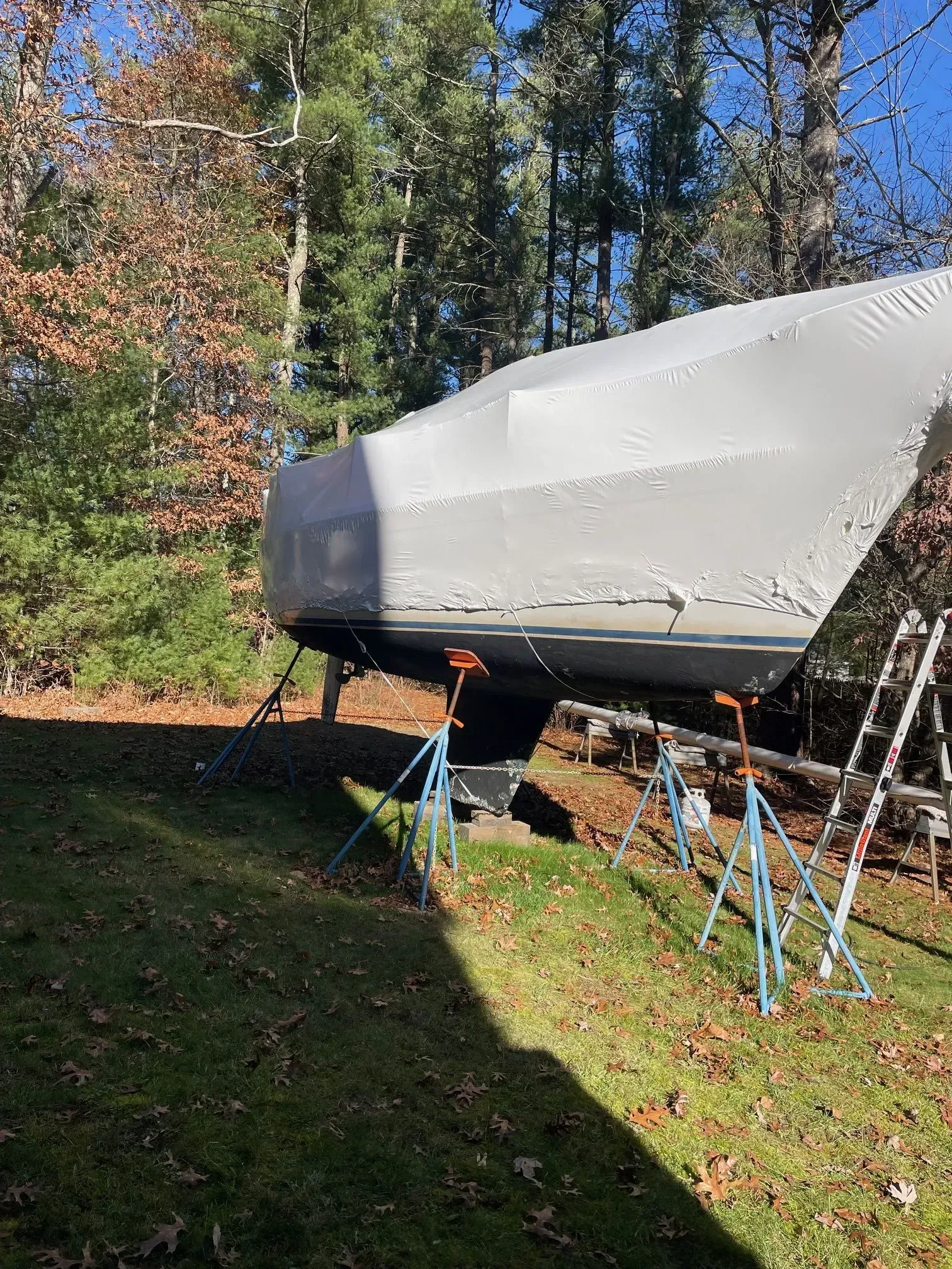 A boat covered in a white tarp supported by blue jack stands on a grassy lawn with trees in the background.