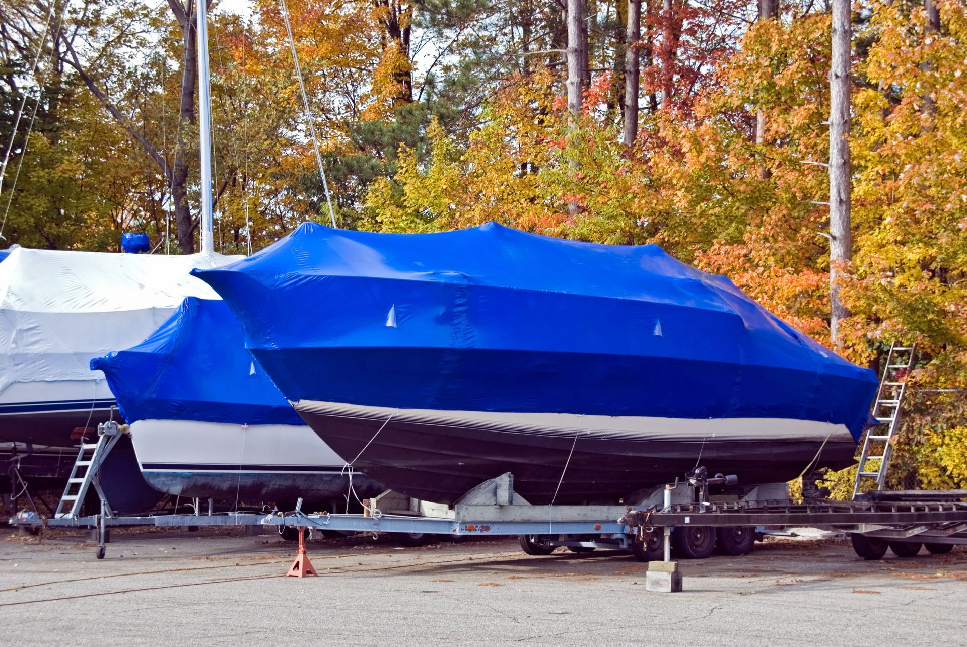 Boats on trailers, covered in blue and white tarps, in a yard with fall foliage.