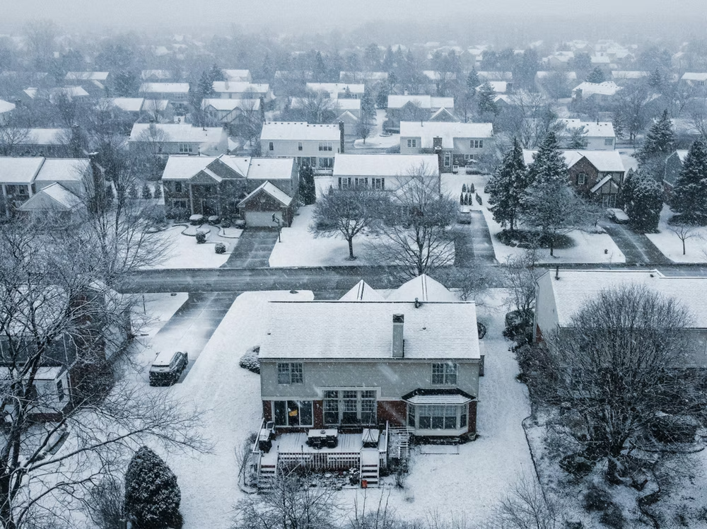 An aerial view of a suburban neighborhood blanketed in snow, featuring houses, snow-covered lawns, and bare trees.