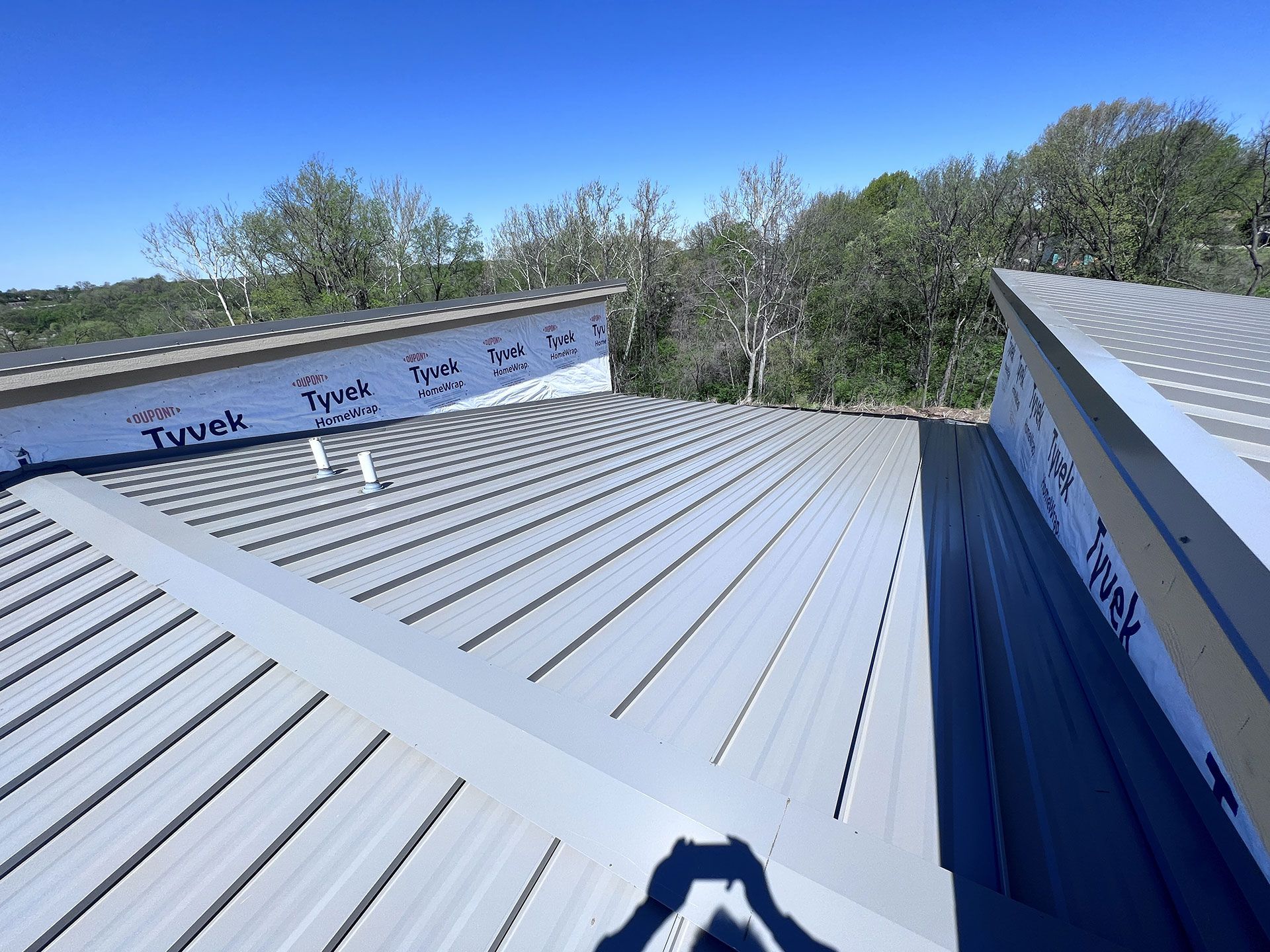 An elevated view of a gray metal roof under construction with Tyvek-wrapped wall sections and trees in the background.