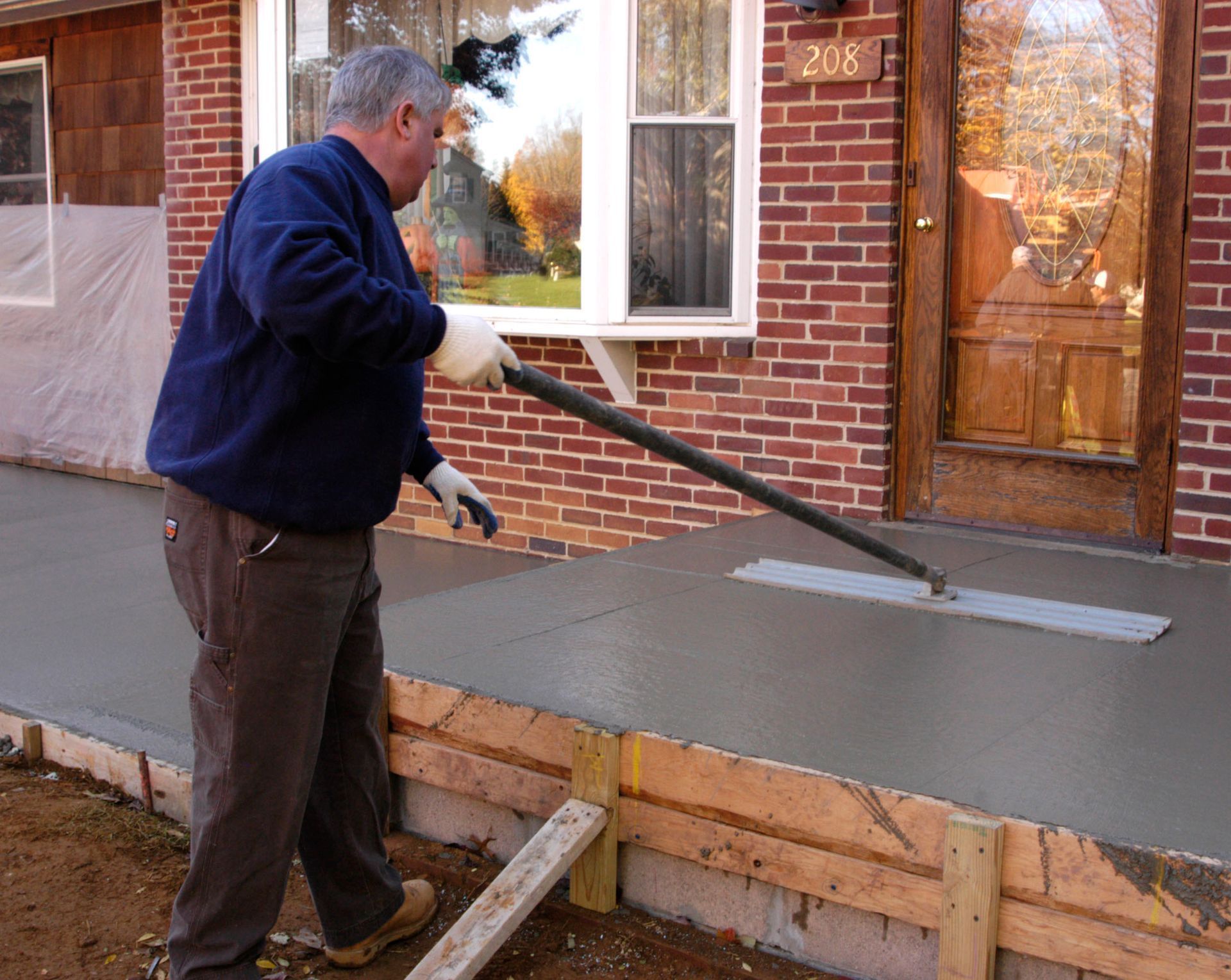 Man smoothing wet concrete on a porch with a long-handled tool. Brick house in background.