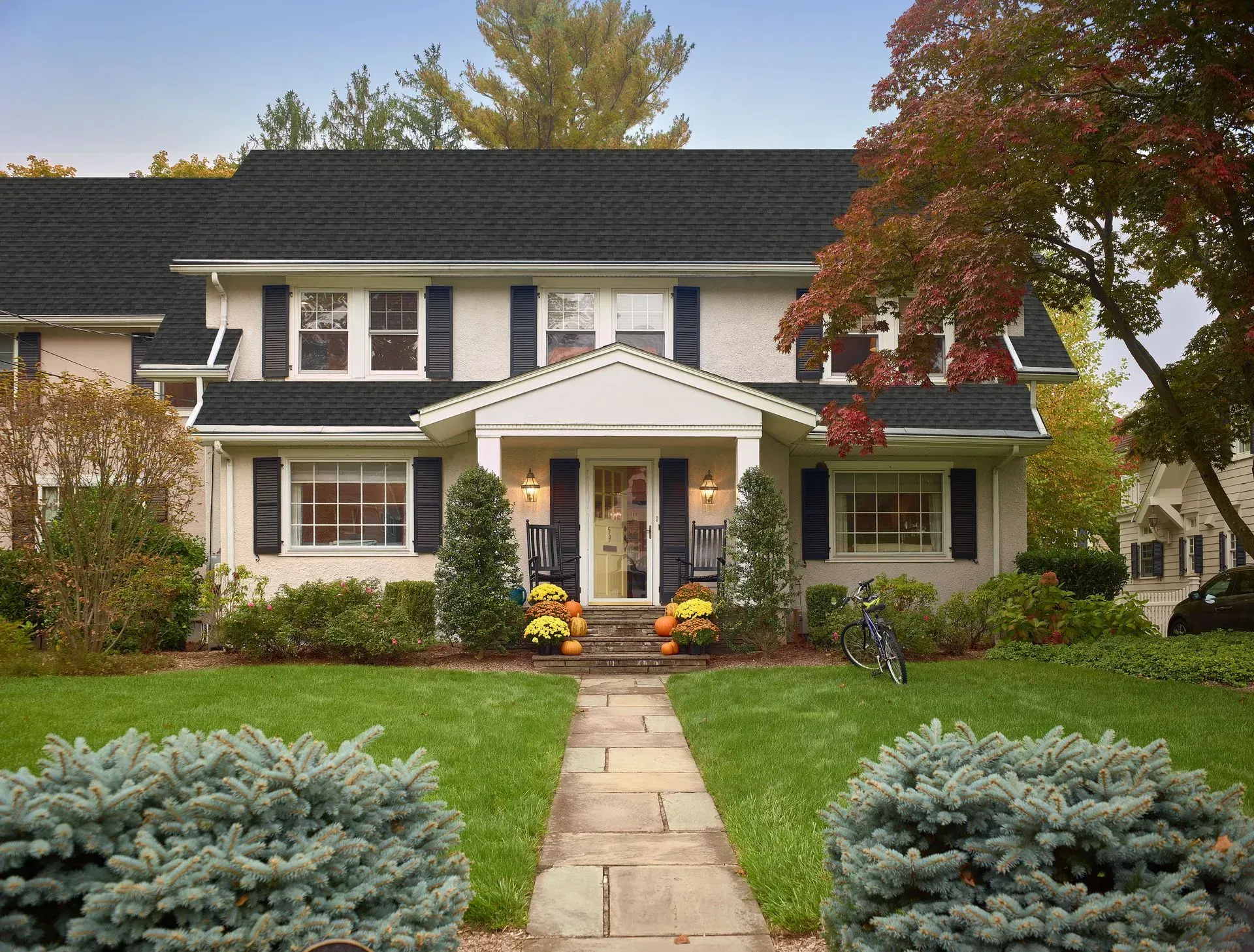 Two-story beige house with black shutters and a stone walkway lined by shrubs. Autumn leaves in the trees.