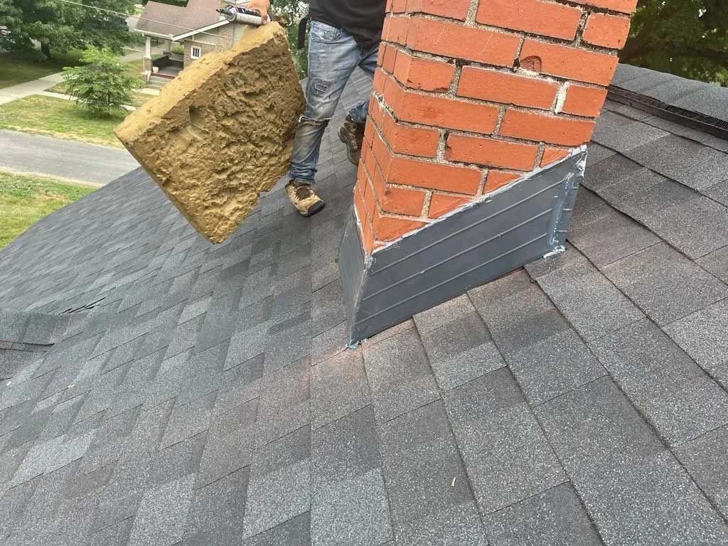 Person on a roof installing insulation near a brick chimney; grey shingles, green grass.