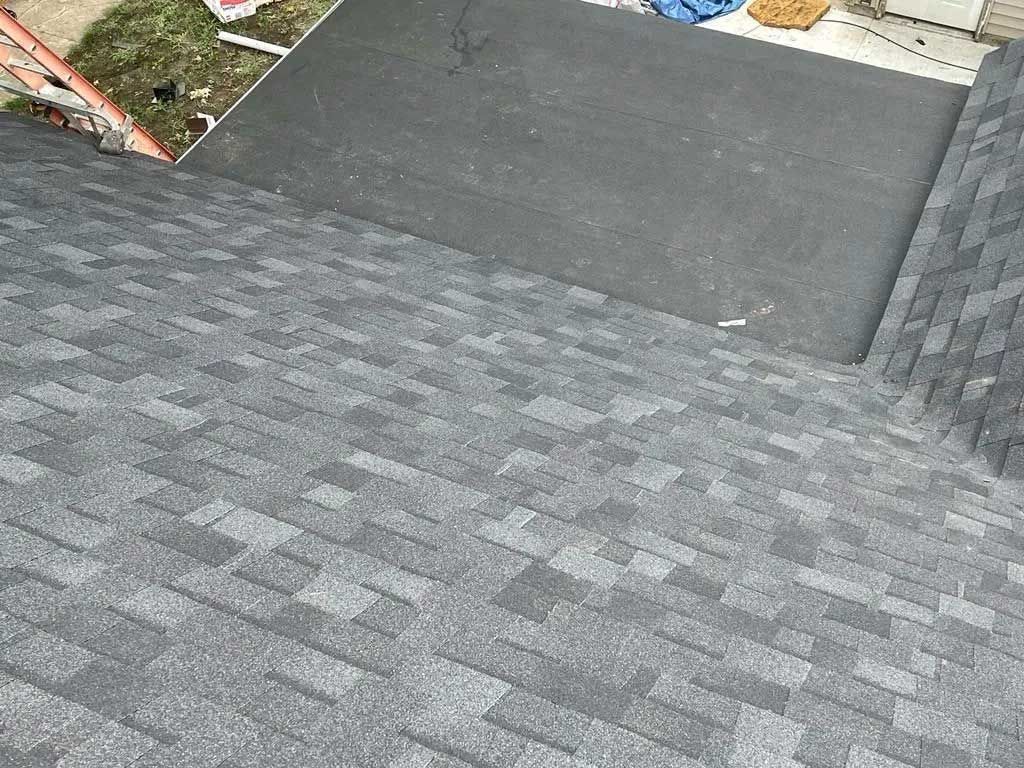 Gray asphalt shingle roof with a dark gray flat surface and part of a lawn and ladder.