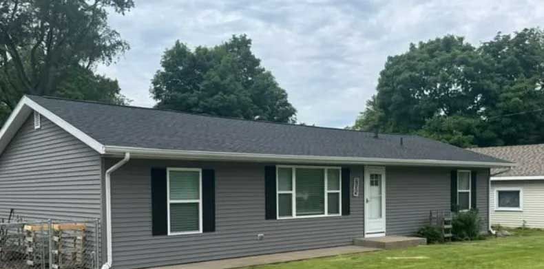Gray house with black shutters, dark roof, and white trim; green lawn and trees in the background.