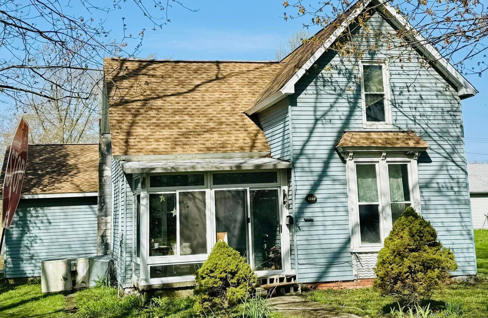 Blue, weathered house with porch and overgrown yard under brown roof, on a sunny day.