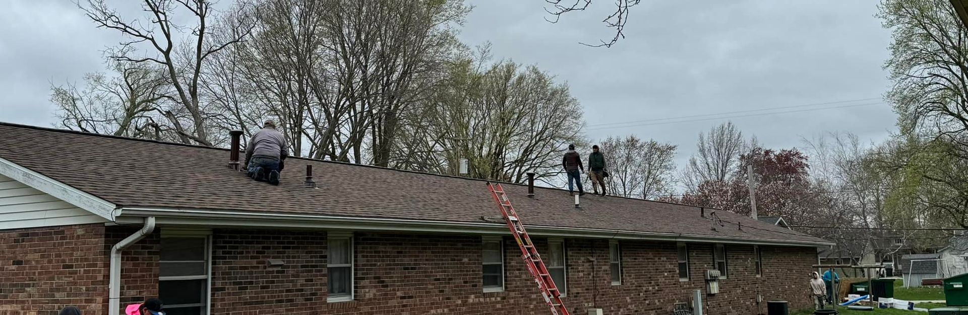 Workers on a brown shingled roof on a brick building, with a ladder, under an overcast sky.