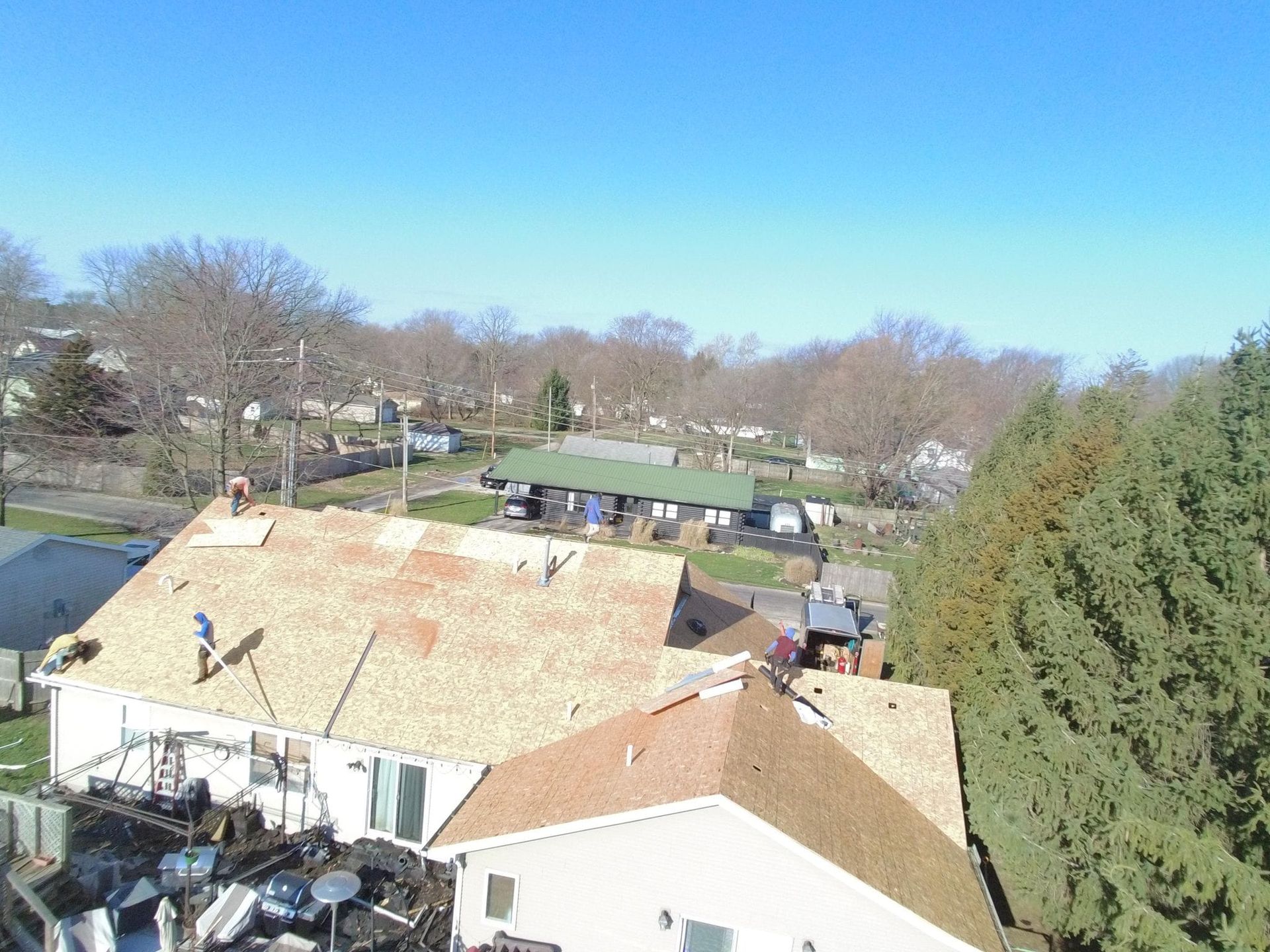 Roofers working on a house roof. Brown shingles, blue sky, and surrounding houses.