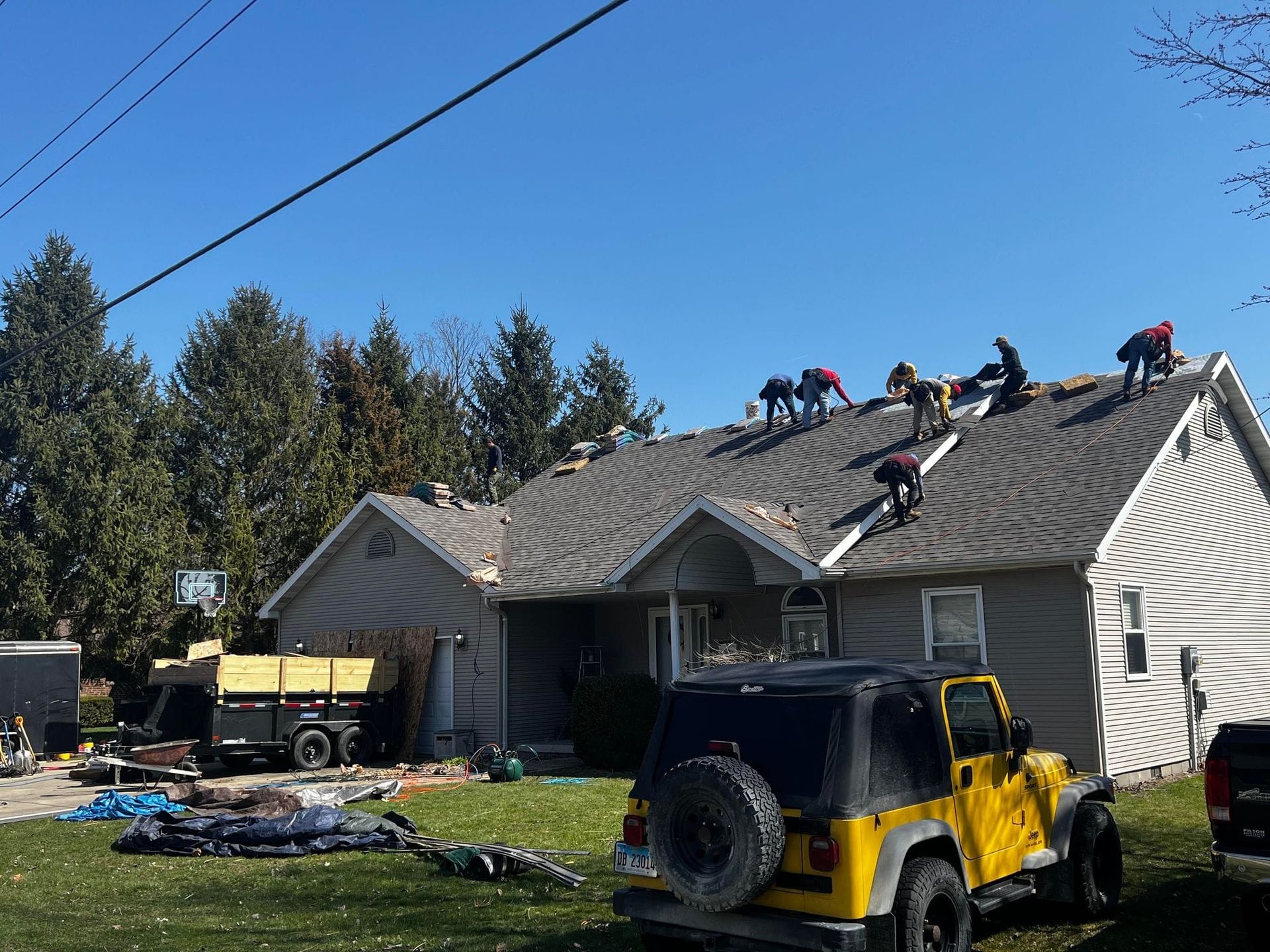 Workers replacing roof shingles on a house under a bright blue sky, with a yellow Jeep in the foreground.