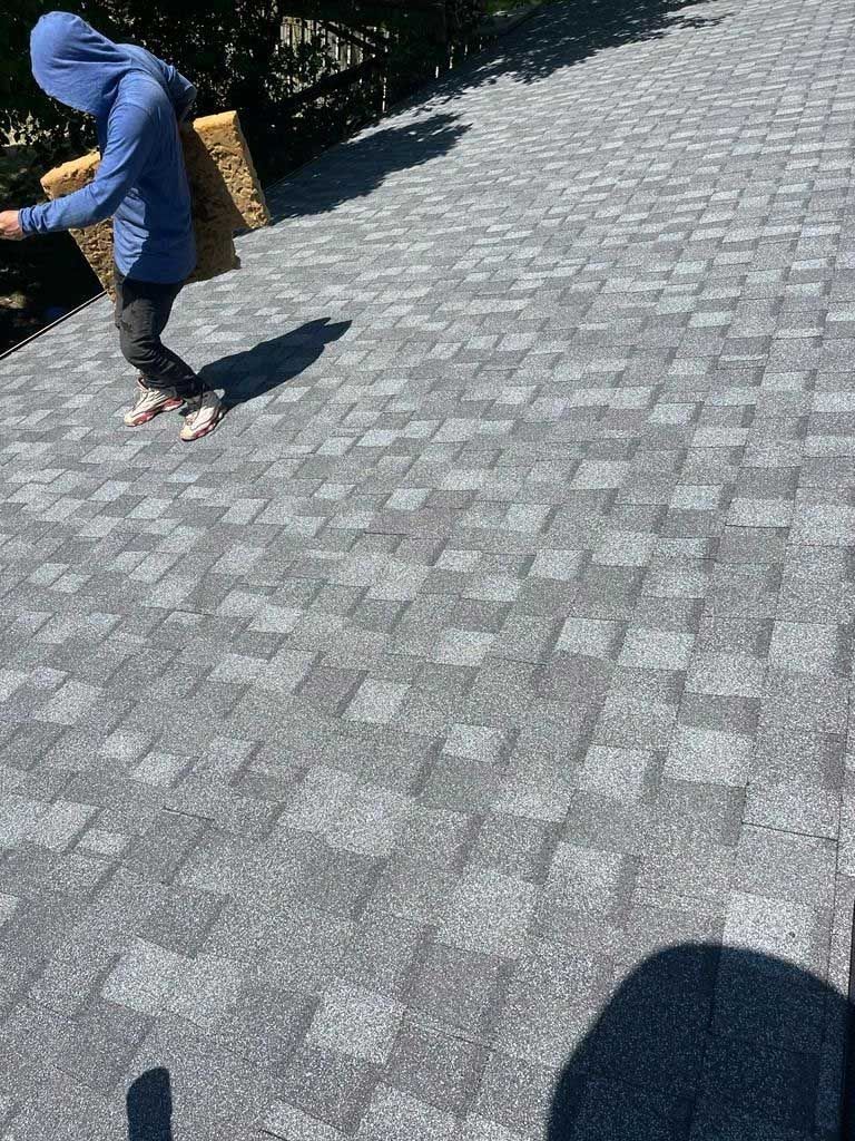 Person in hoodie on a gray shingled roof, carrying a square item, with shadow.