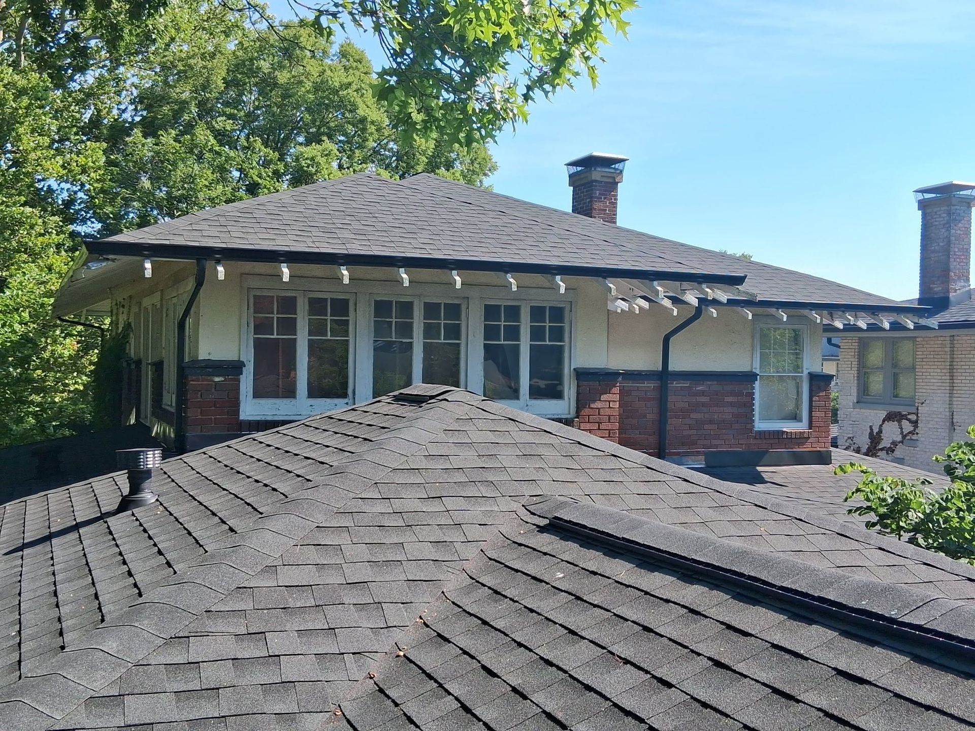 Rooftop view of a brick house with gray shingles, white-trimmed windows, and trees against a blue sky