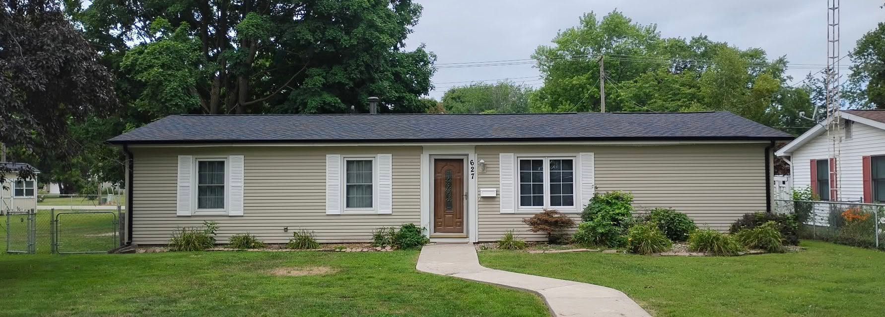Single-story beige house with white trim, dark roof, front walkway, and green lawn under cloudy skies