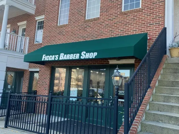 "Figga's Barber Shop, a brick building with a green awning. The shop has a black fence and stairs."