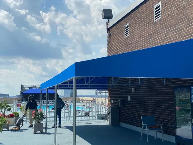 Blue awning on a rooftop, with a pool and people near a brick building.