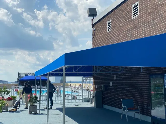 Blue awning on a rooftop, with a pool and people near a brick building.