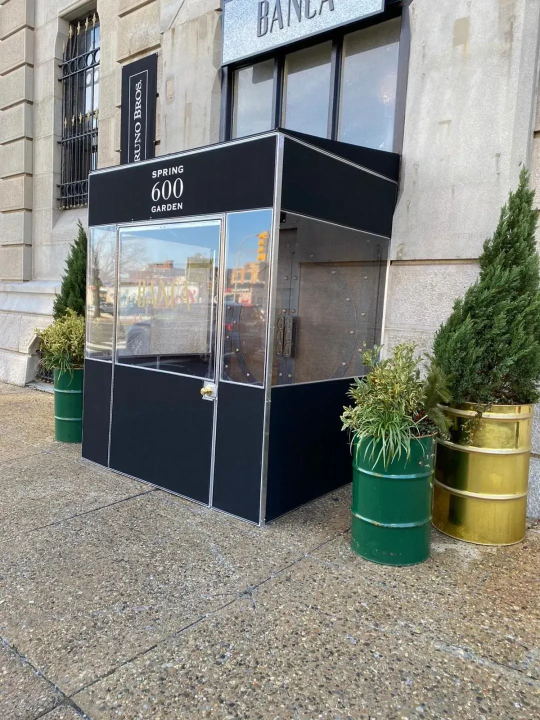 Black and silver kiosk with glass windows outside a building, flanked by green and gold planters.