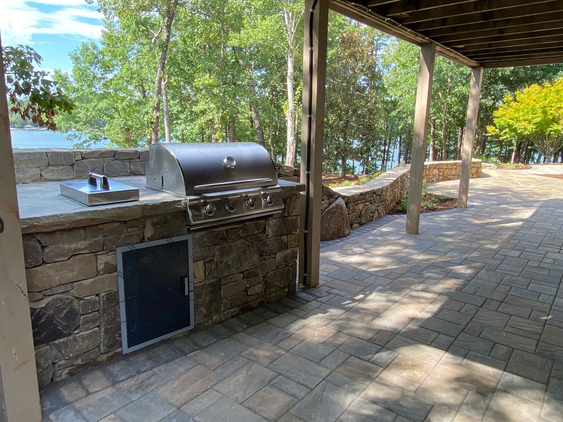 A grill is sitting under a pergola on a patio next to a lake.