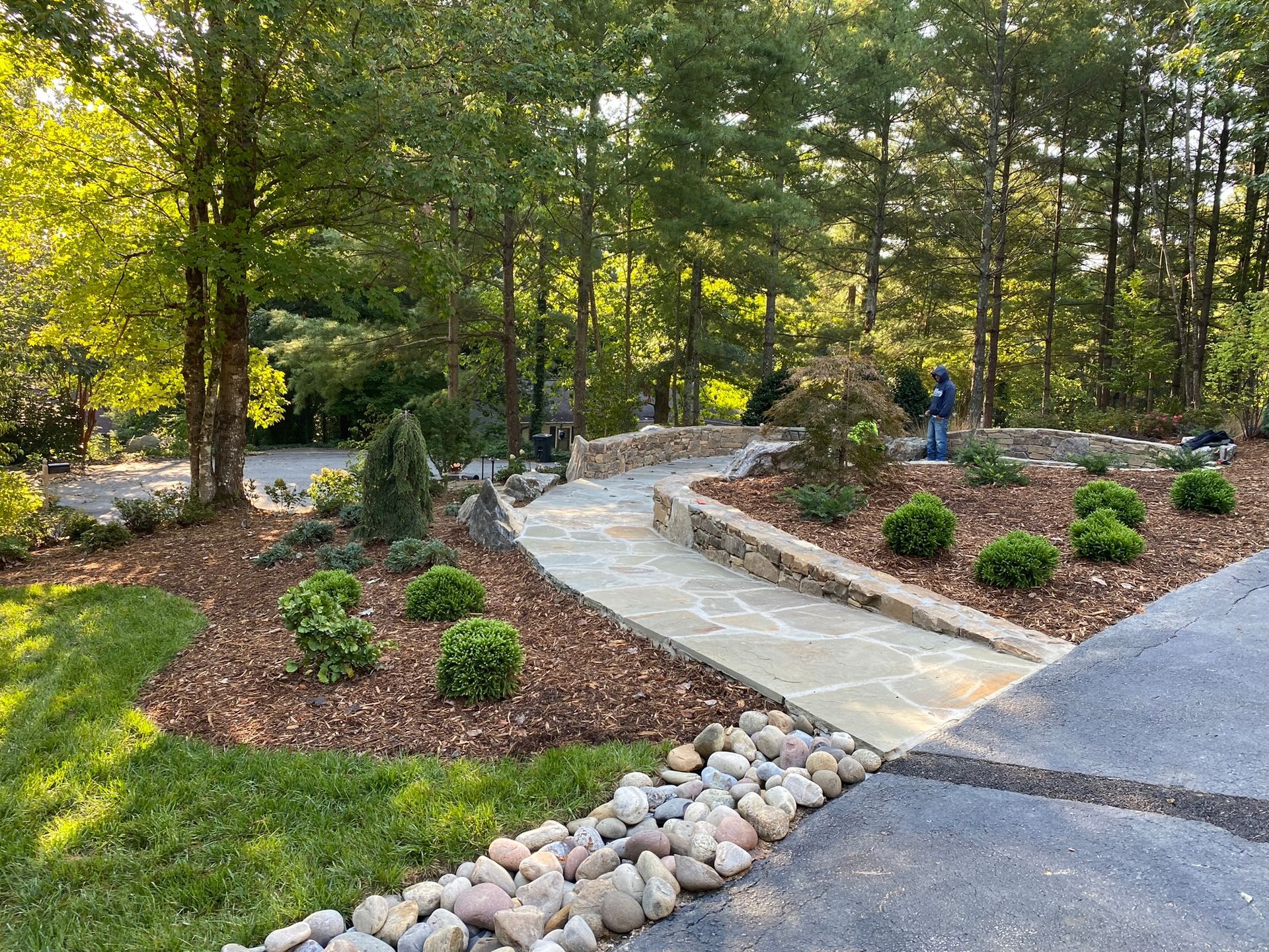 A stone walkway leading to a driveway surrounded by trees and rocks.
