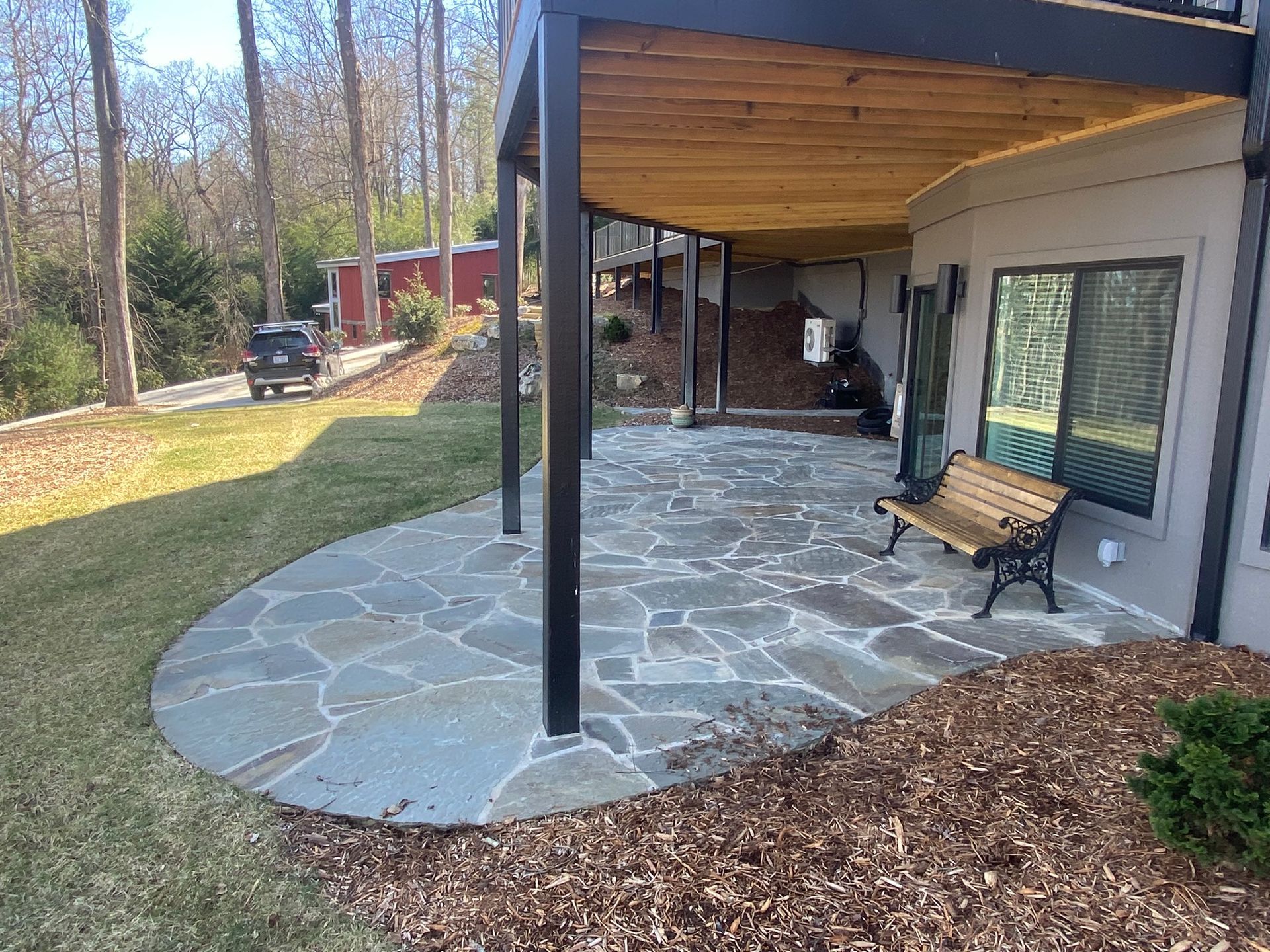 A stone patio with a bench under a wooden pergola in front of a house.