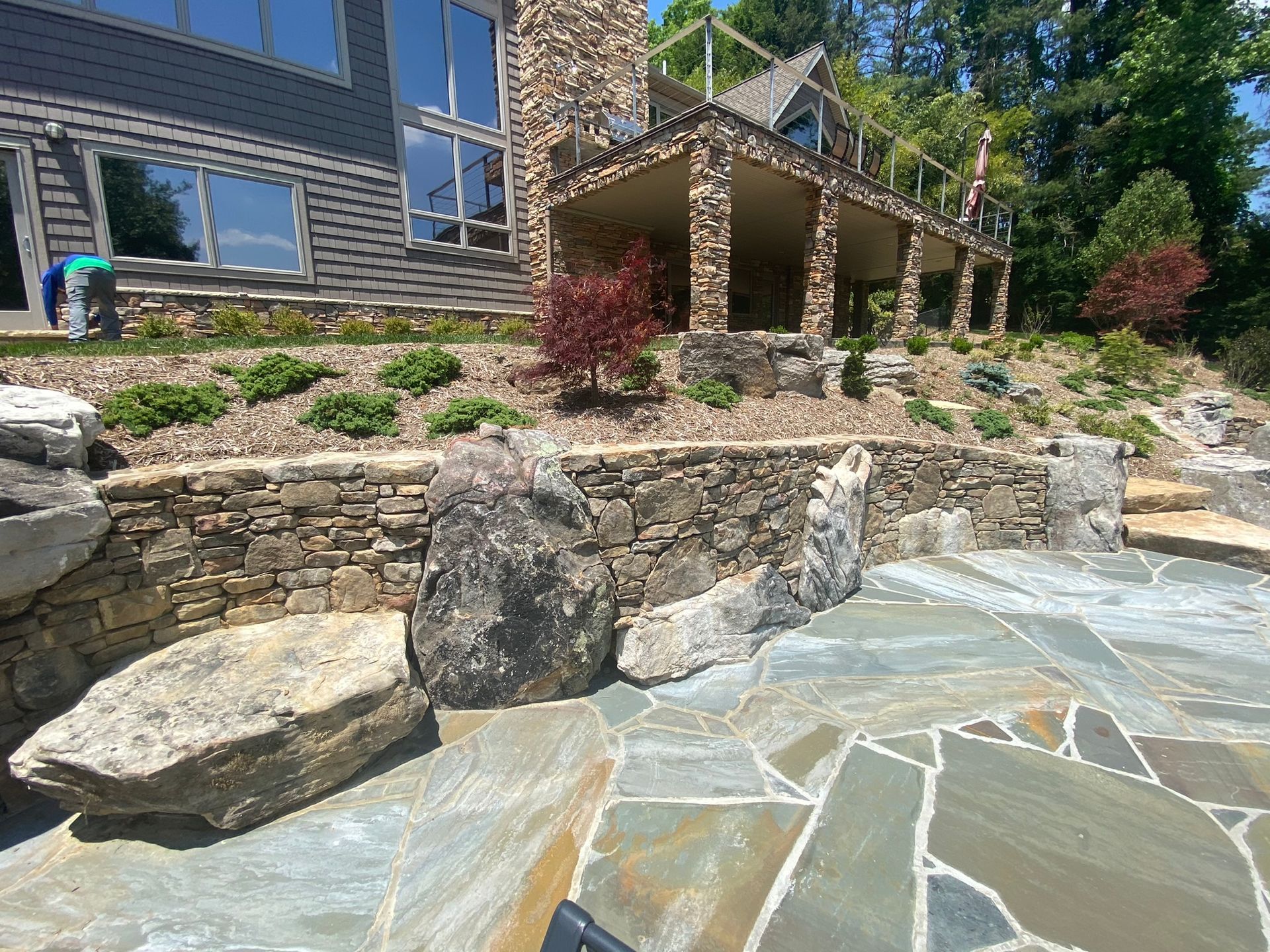 A large stone wall surrounds a swimming pool in front of a large house.