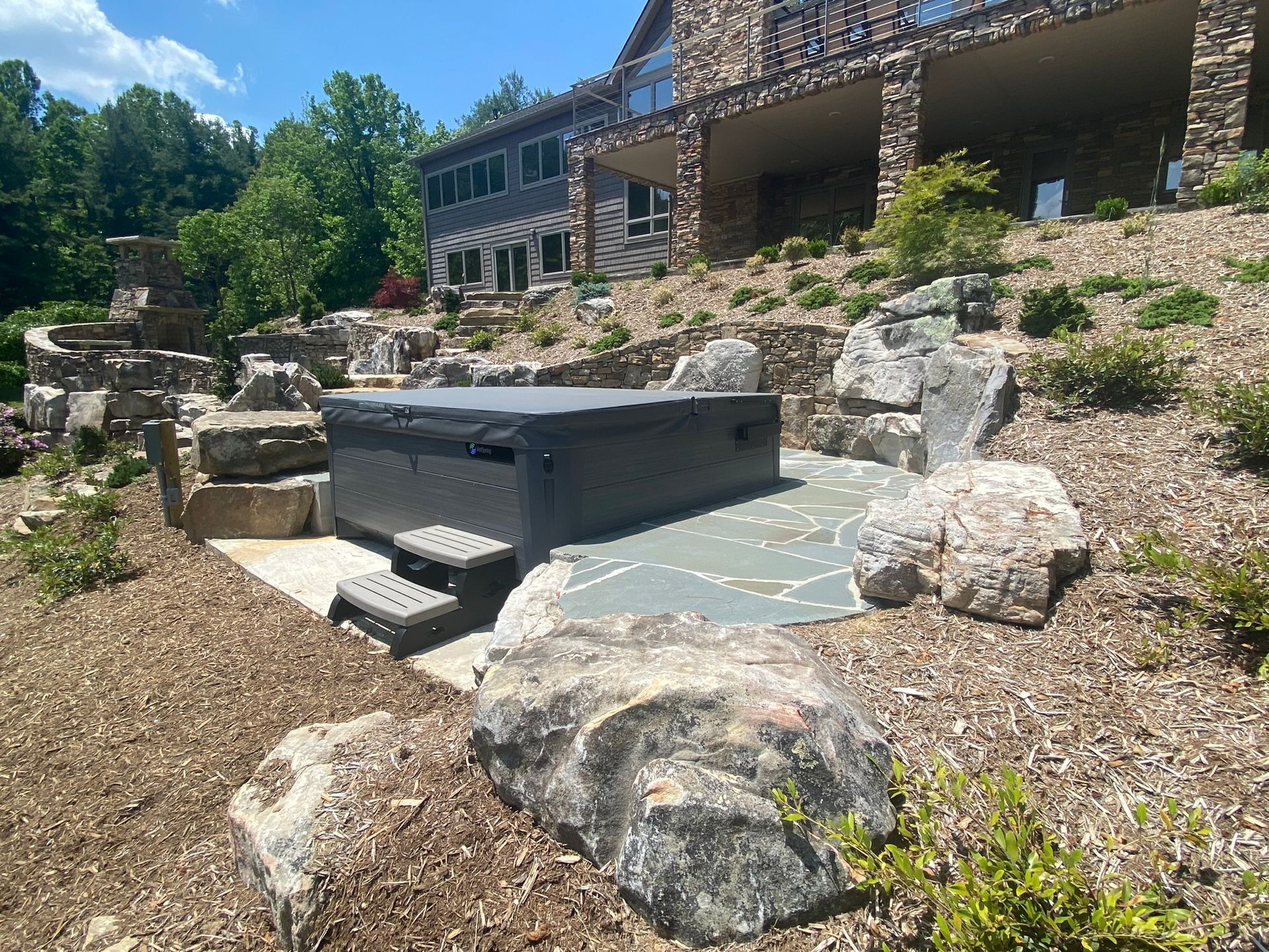 A hot tub is sitting in the middle of a rocky area in front of a large house.