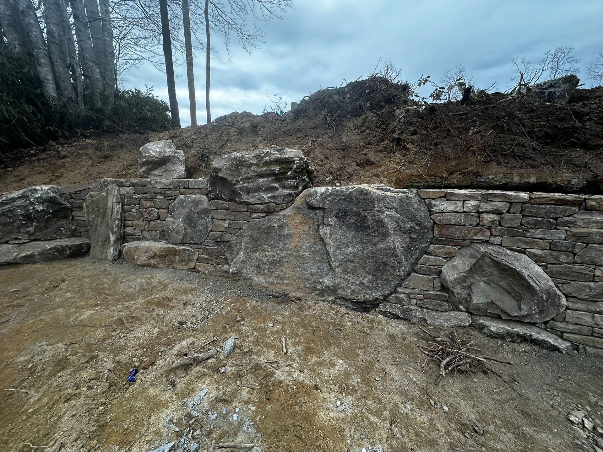 A large rock is sitting in the middle of a dirt field next to a stone wall.