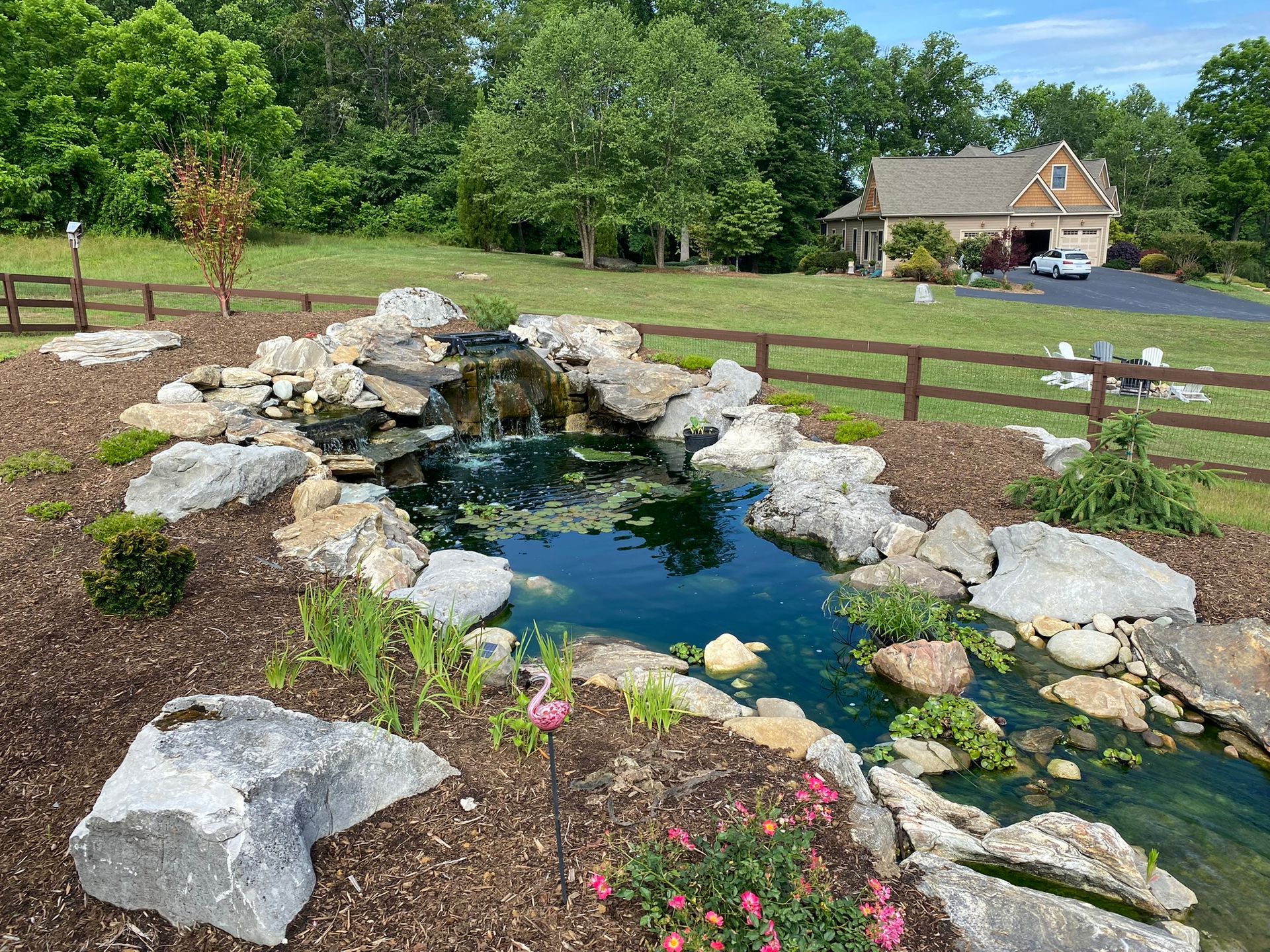 A pond with a waterfall in the middle of a yard in front of a house.
