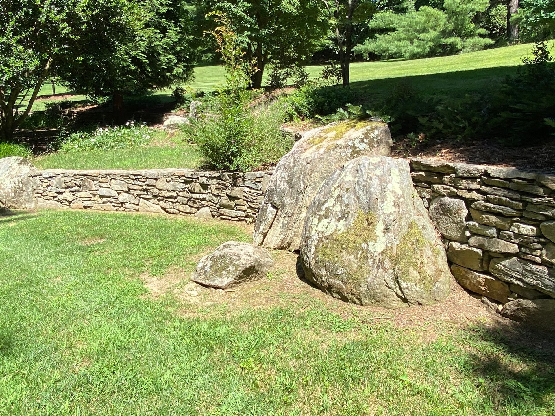 A large rock is sitting in the middle of a lush green field next to a stone wall.