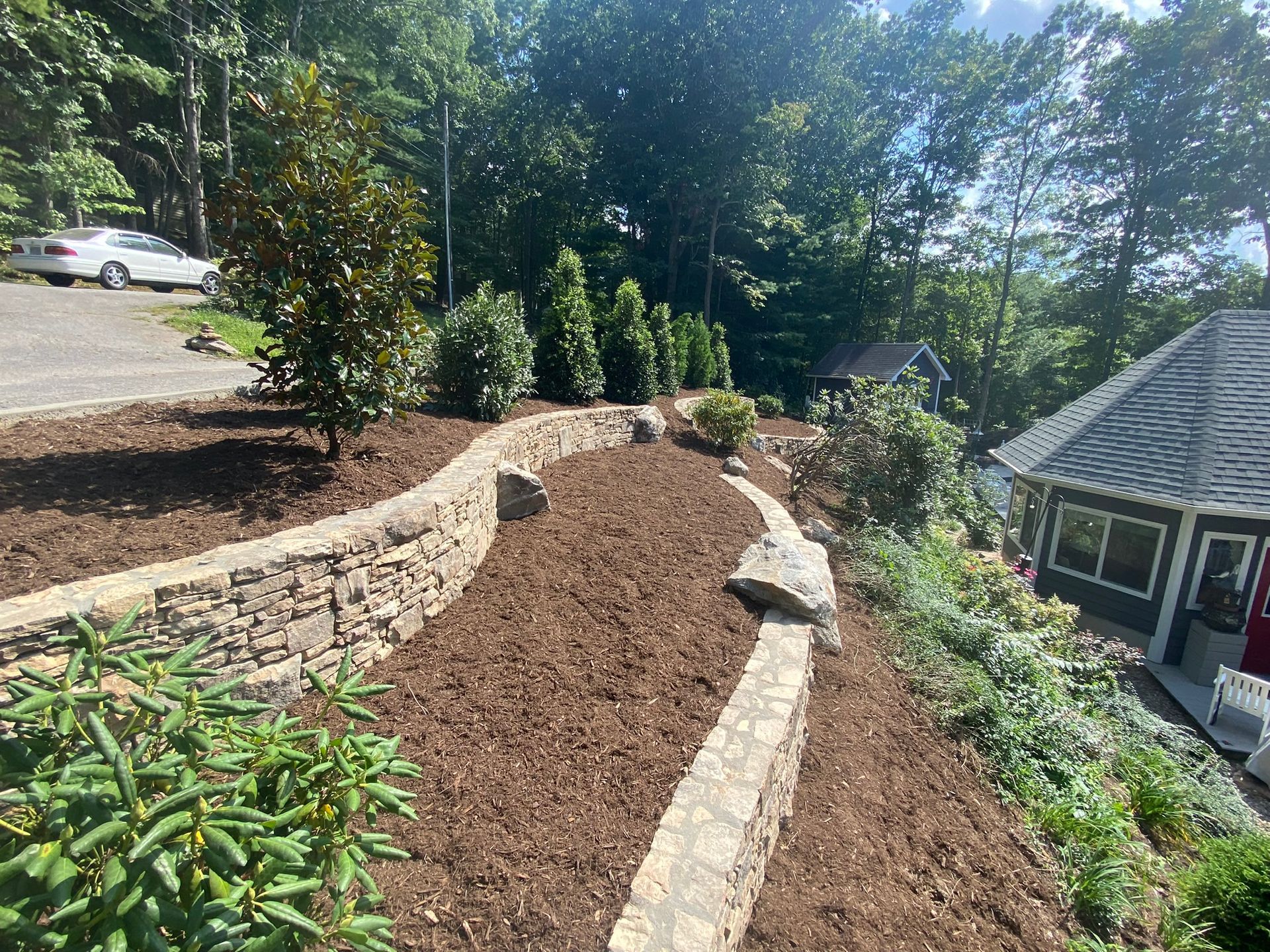 A stone wall is surrounded by mulch and trees in front of a house.