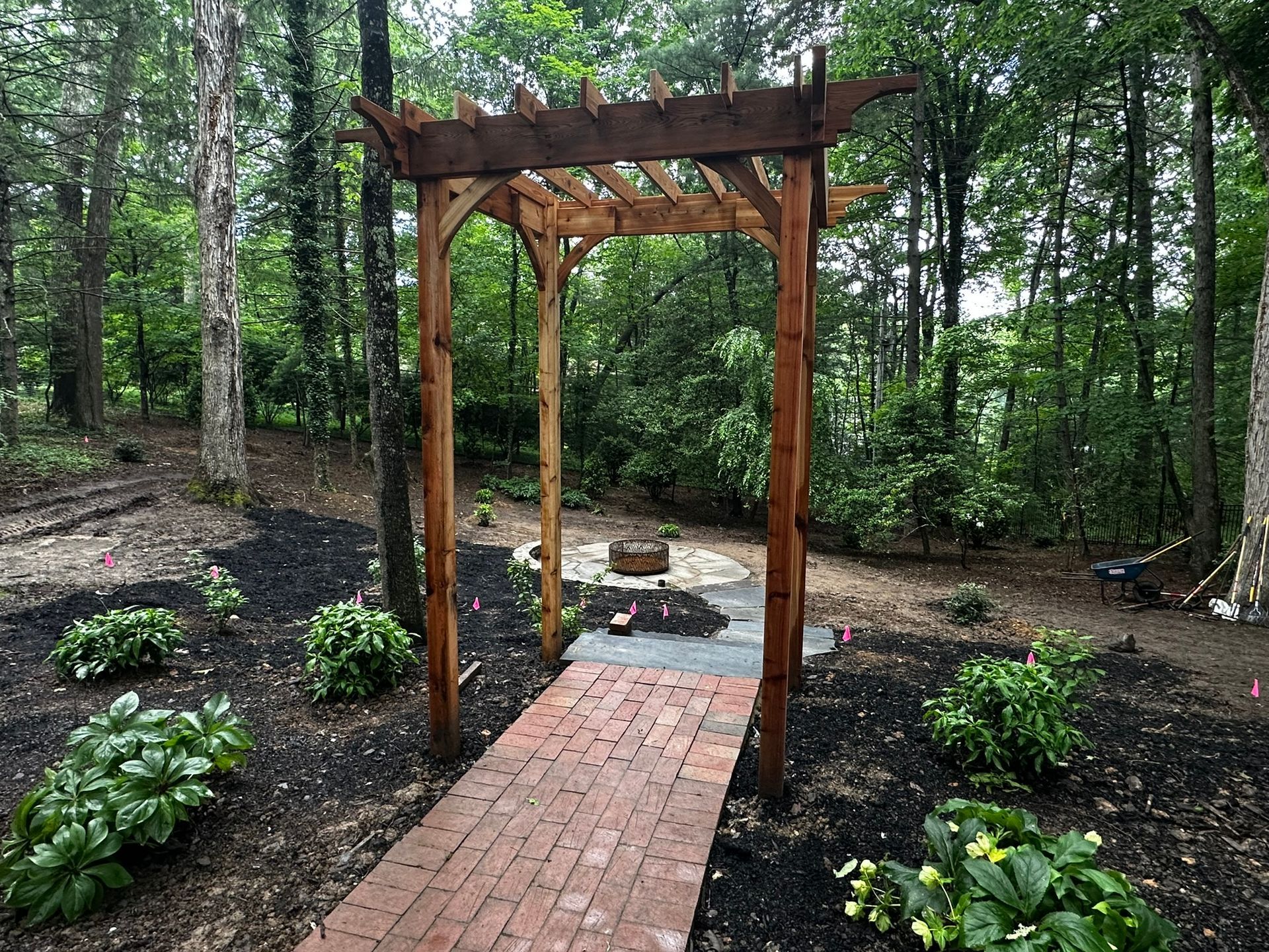 A wooden pergola with a brick walkway leading to it in the middle of a forest.