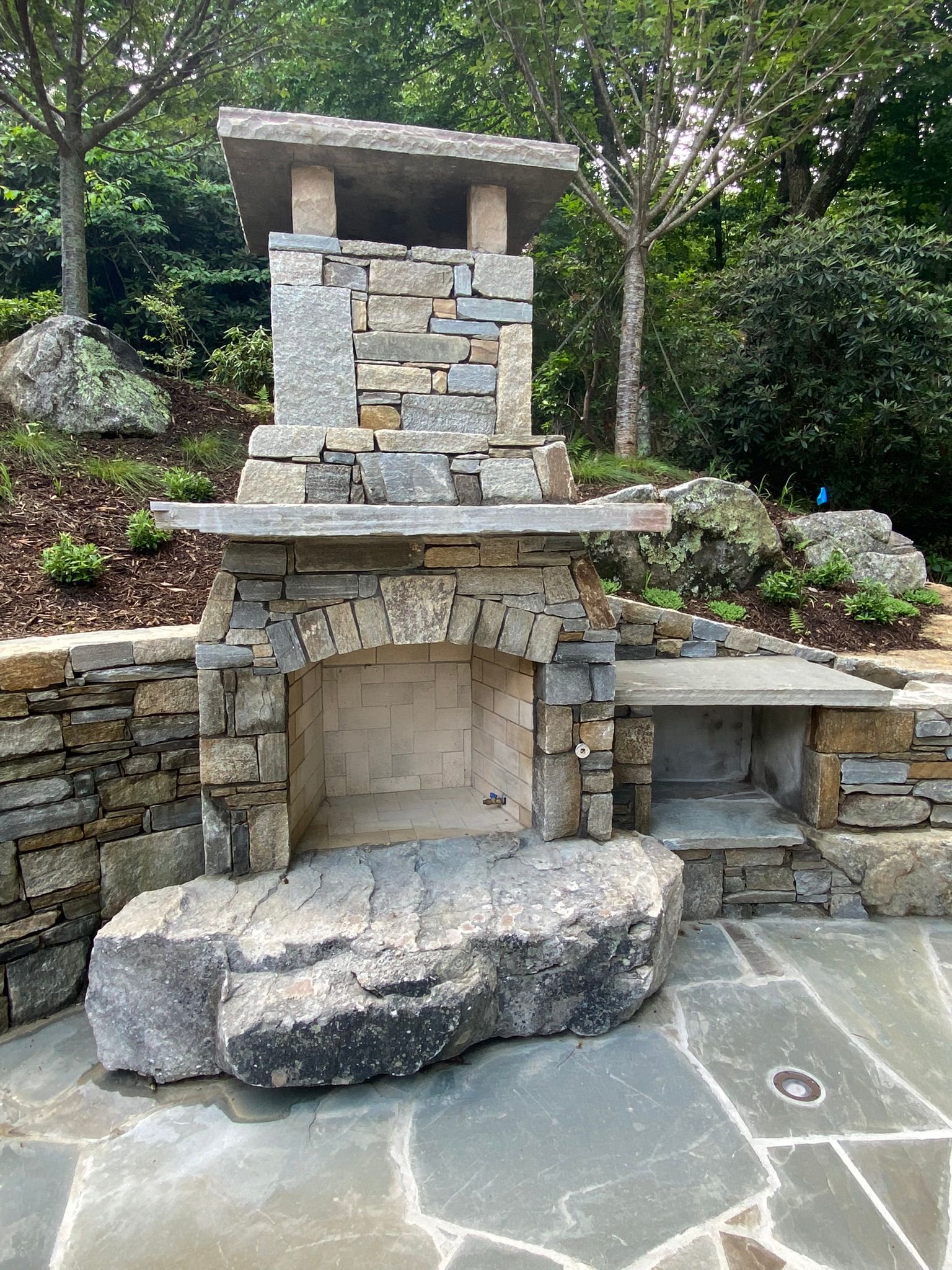 A stone fireplace is sitting on top of a stone patio next to a stone wall.