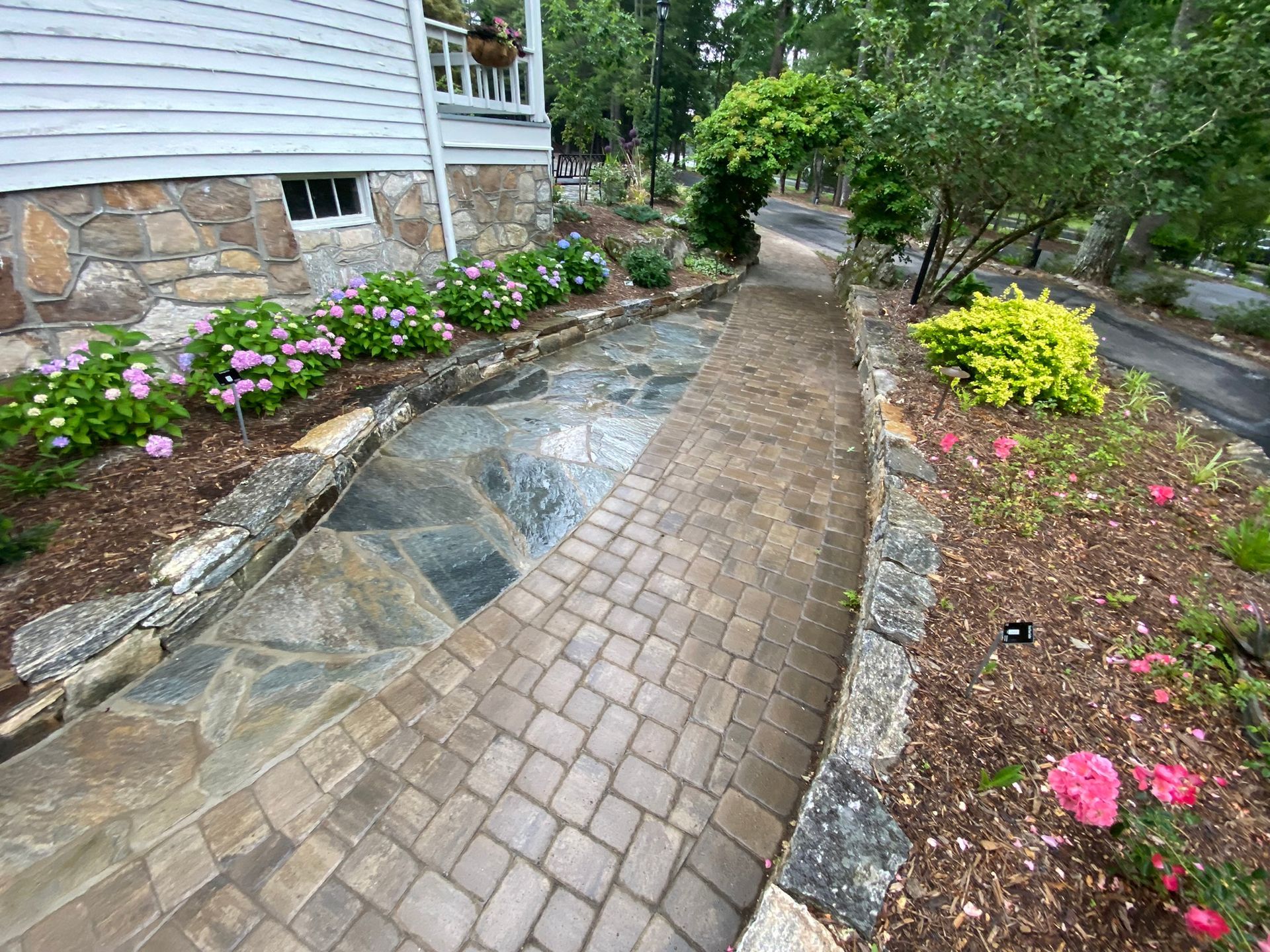 A stone walkway leading to a house surrounded by flowers and bushes.