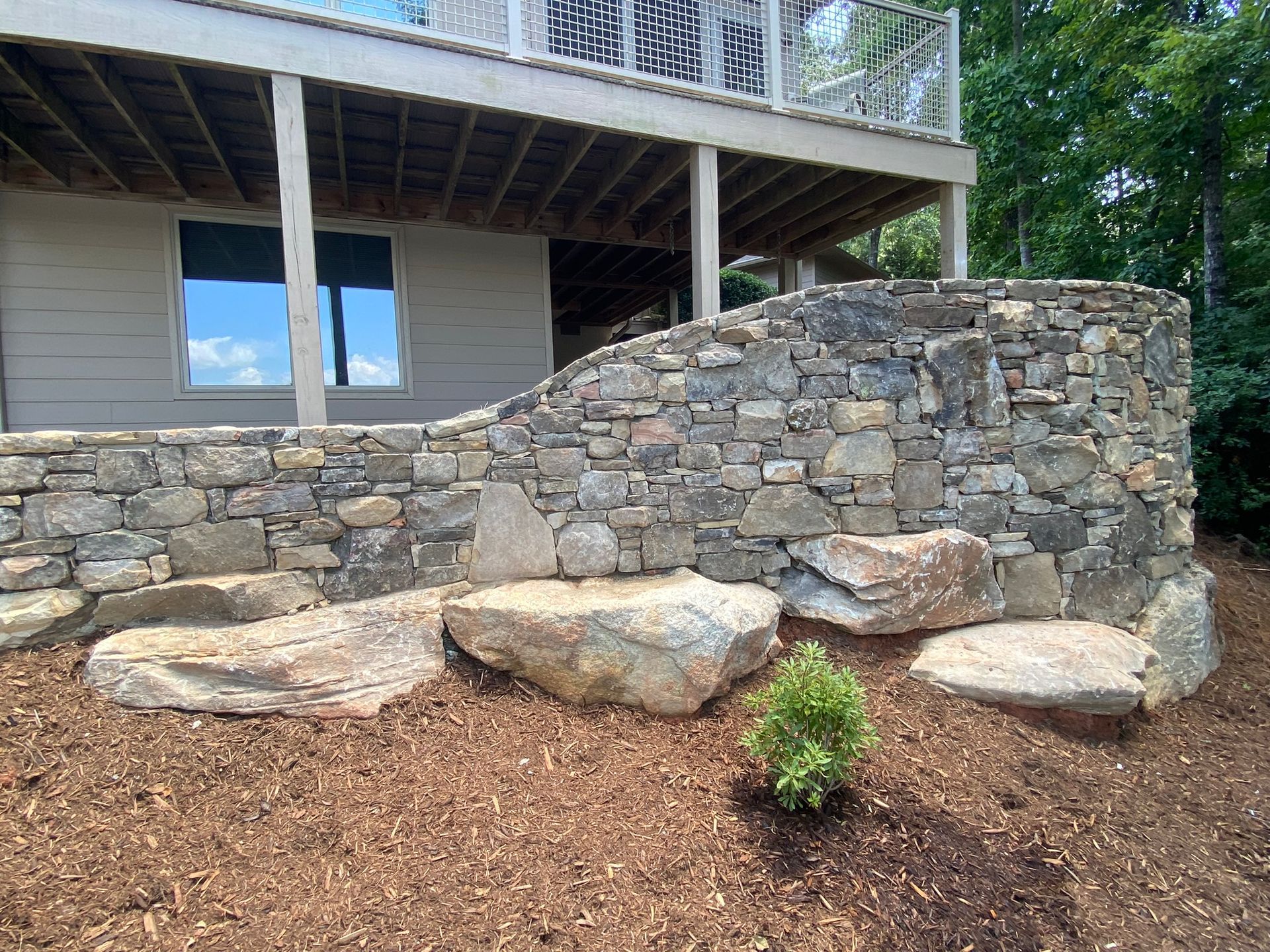 A stone wall is in front of a house with a deck.
