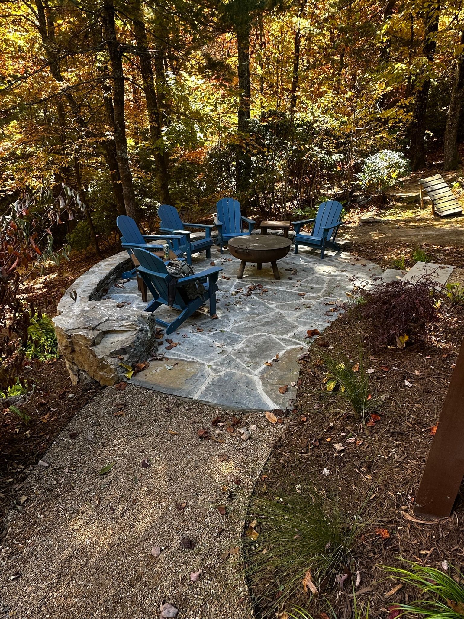 There is a fire pit in the middle of the woods with blue chairs.