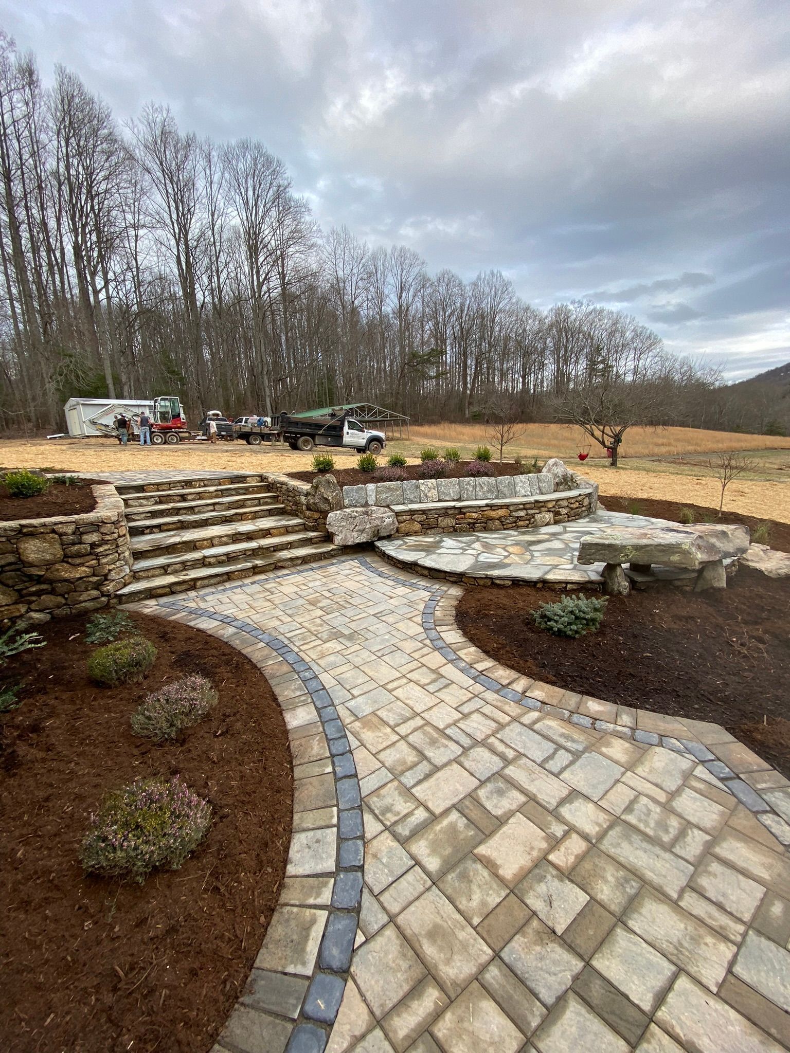 A brick walkway leading to a stone wall and stairs in a backyard.