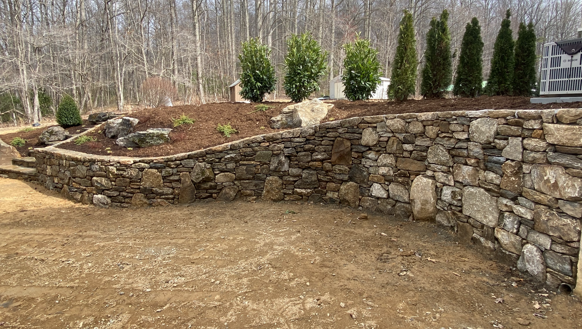 A stone wall is surrounded by trees and dirt in a yard.