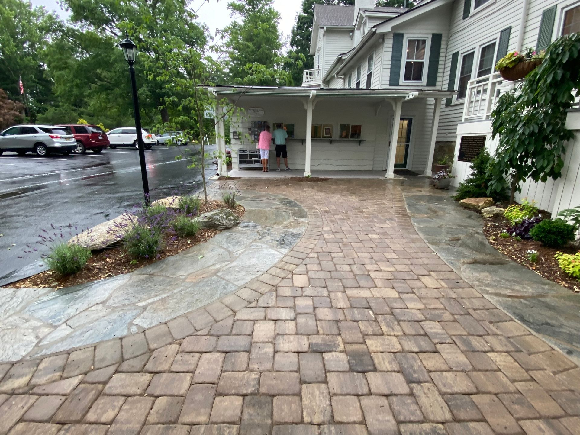 A brick walkway leading to a white house with cars parked in front of it.