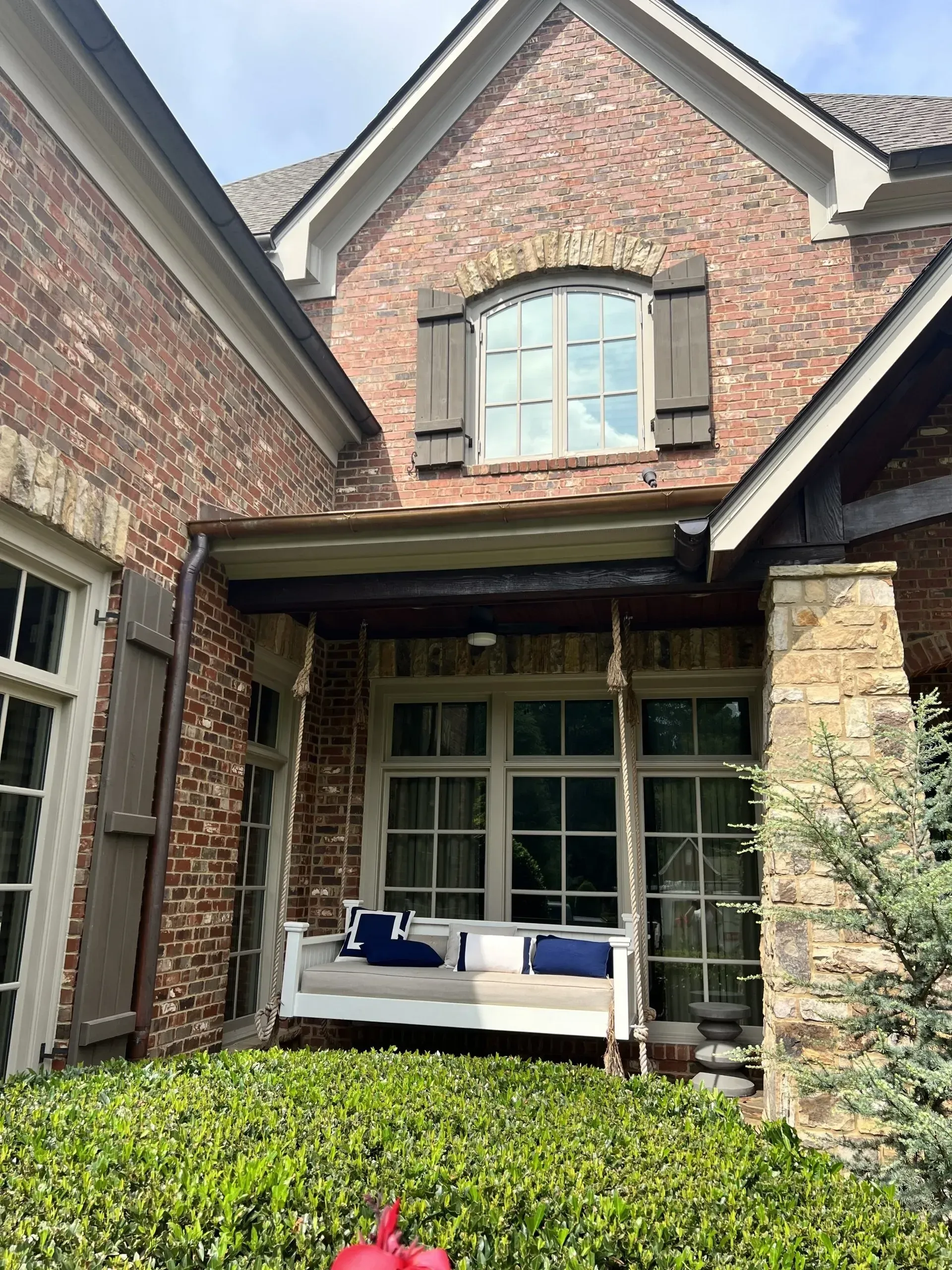 Brick house exterior with swing seat under a porch, surrounded by greenery.