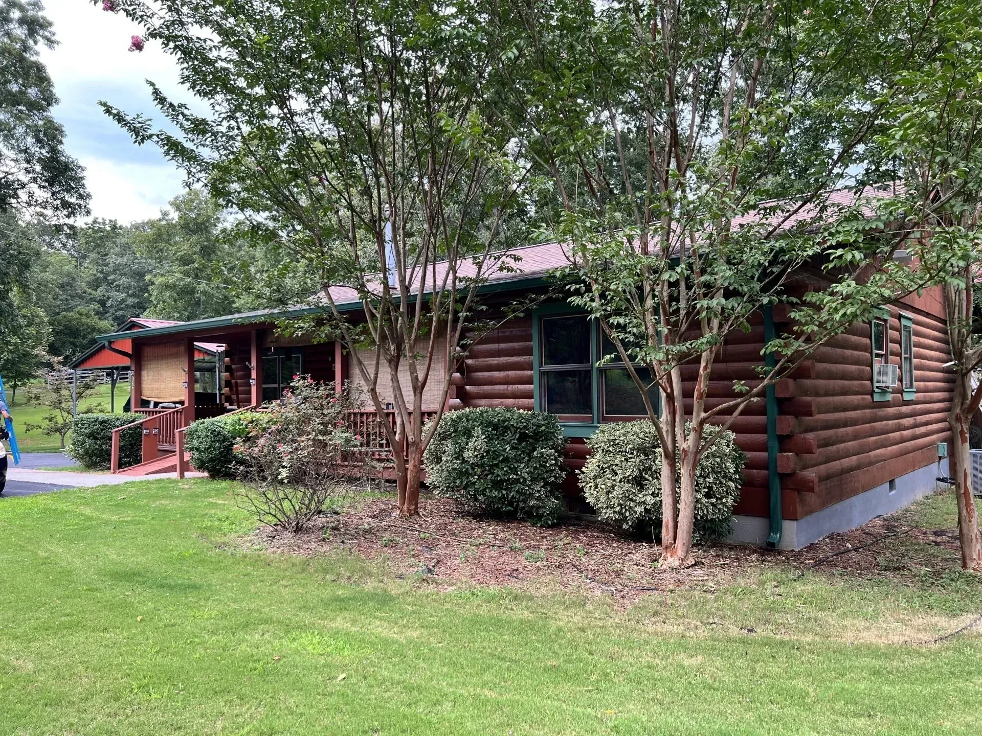 Log cabin with brown logs, green bushes, and lawn; set among trees under a cloudy sky.