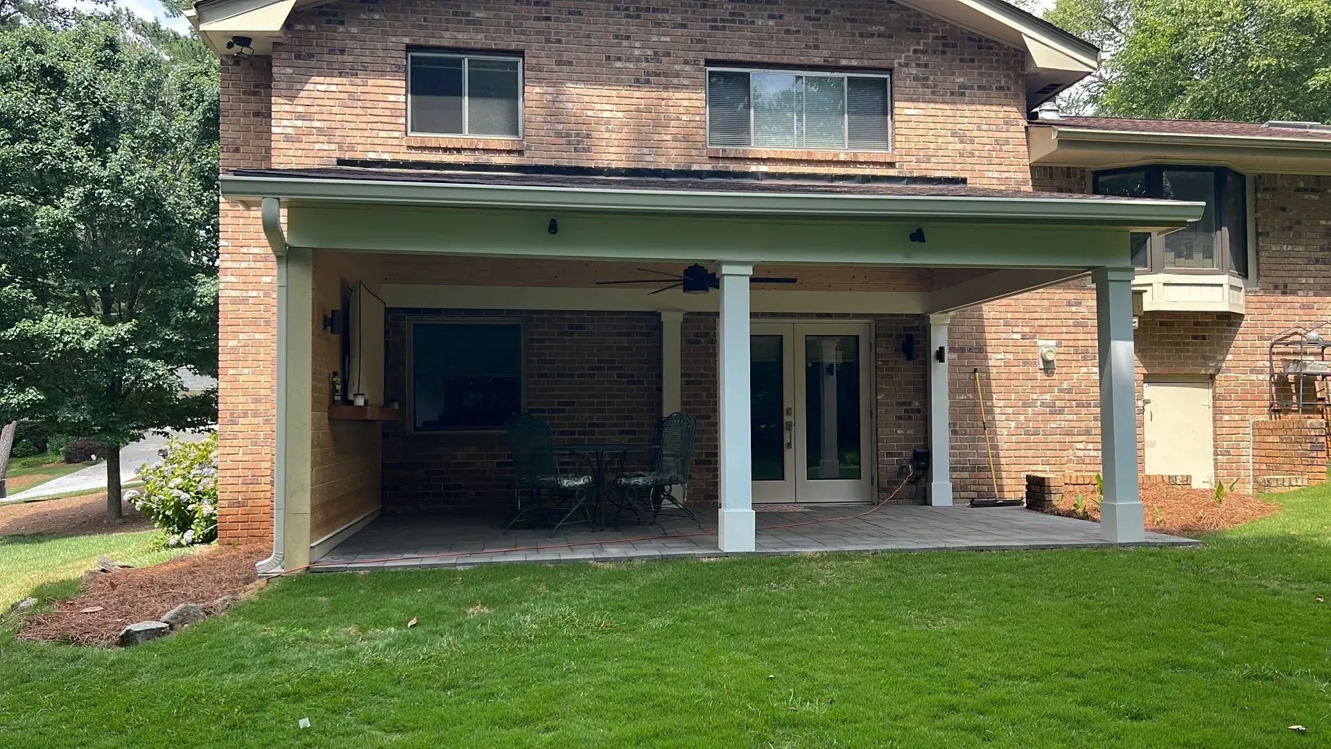 Brick house with a covered patio. White columns support the roof over a concrete patio. Green grass in foreground.