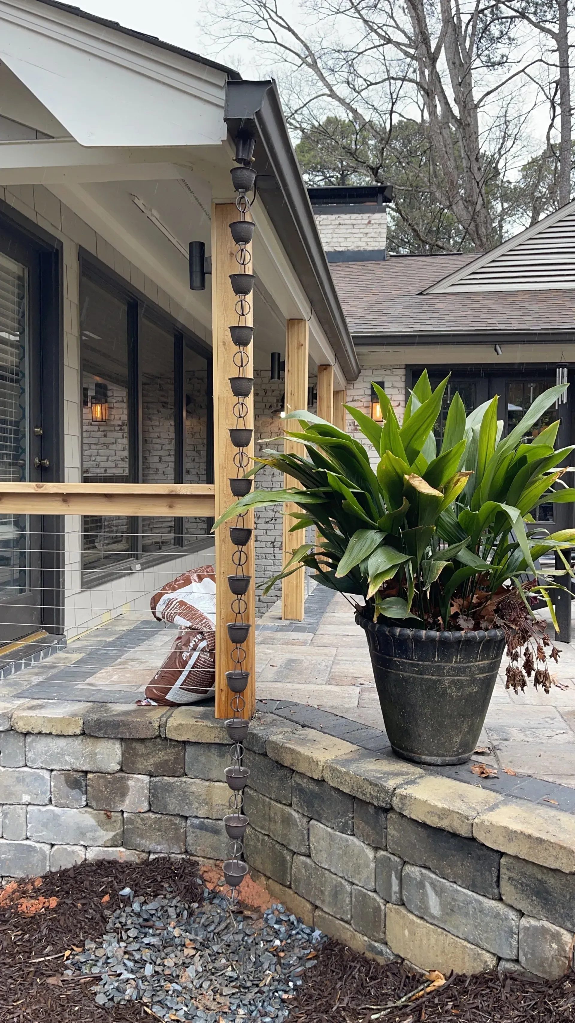 Rain chain installed on a porch post, next to a potted plant on a brick wall.