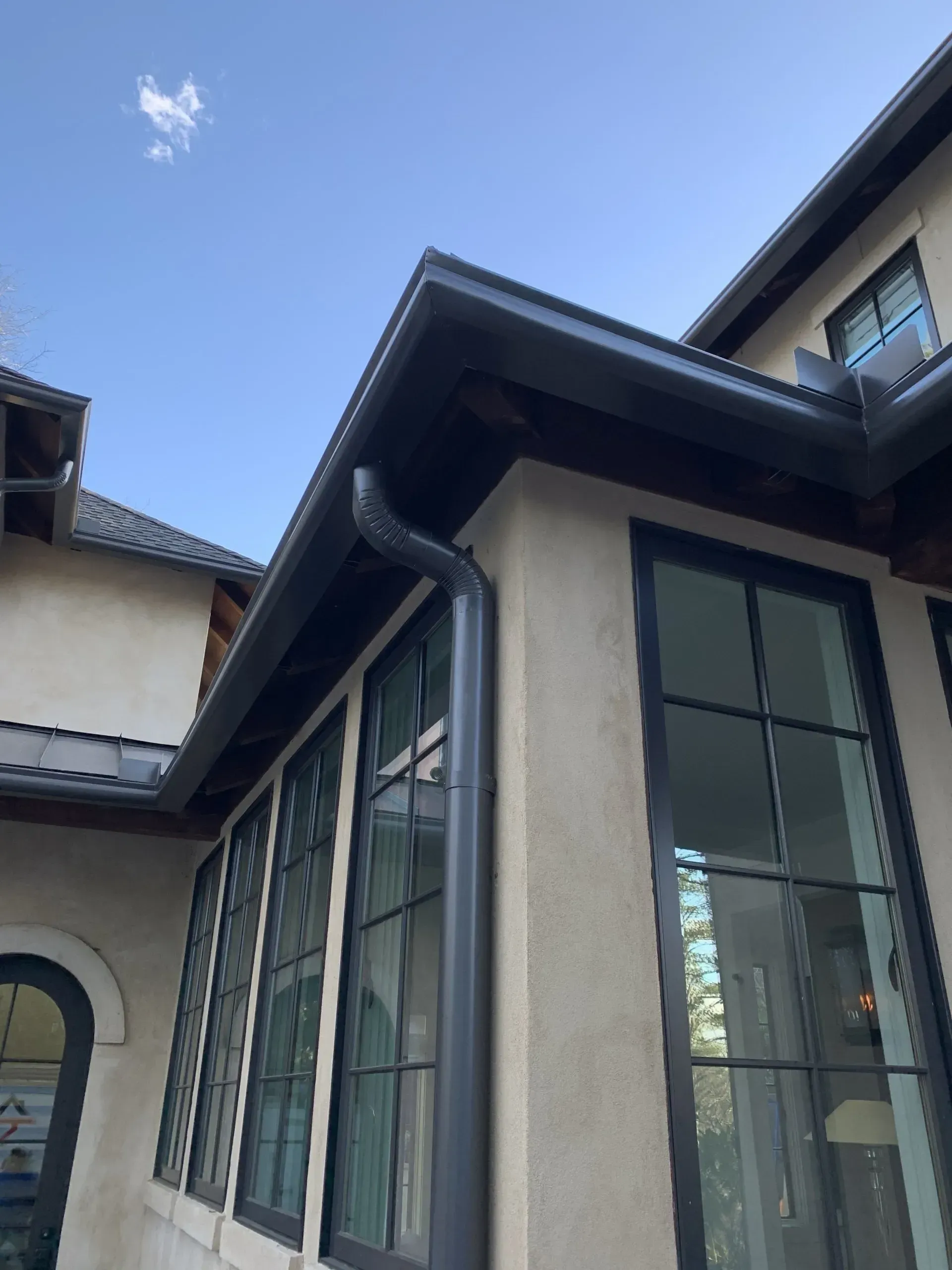 Corner of a house with dark gray gutters and window frames against a light-colored stucco exterior; blue sky.