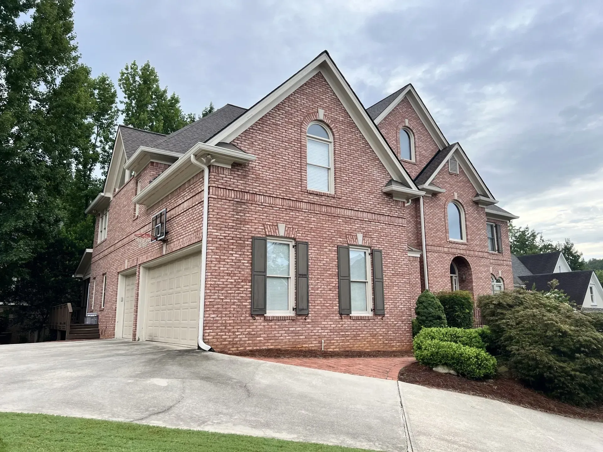 Brick house with two-car garage, brown shutters, and sloping driveway on an overcast day.
