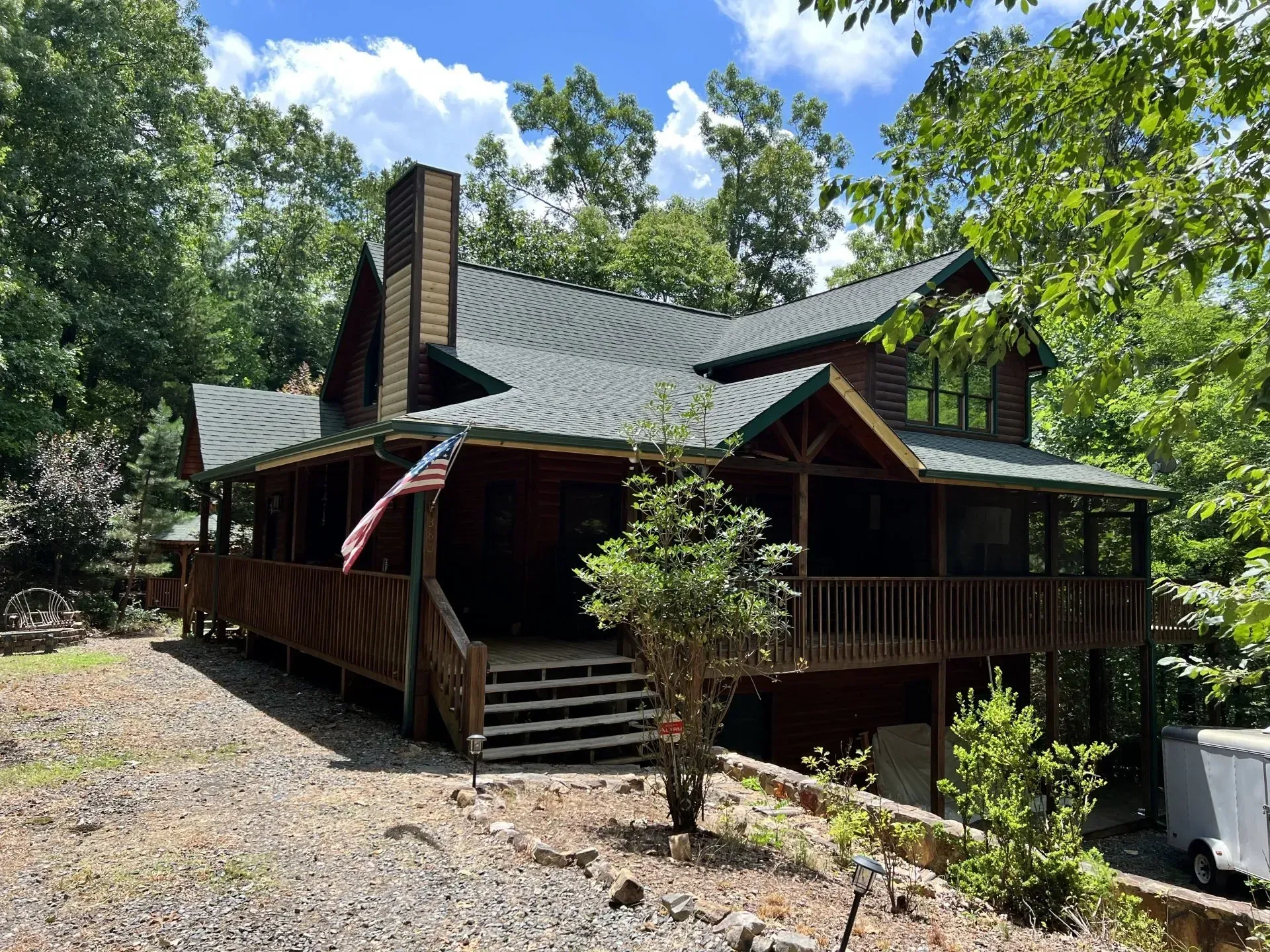 Brown cabin with wrap-around porch, green metal roof, and an American flag. Surrounded by trees.