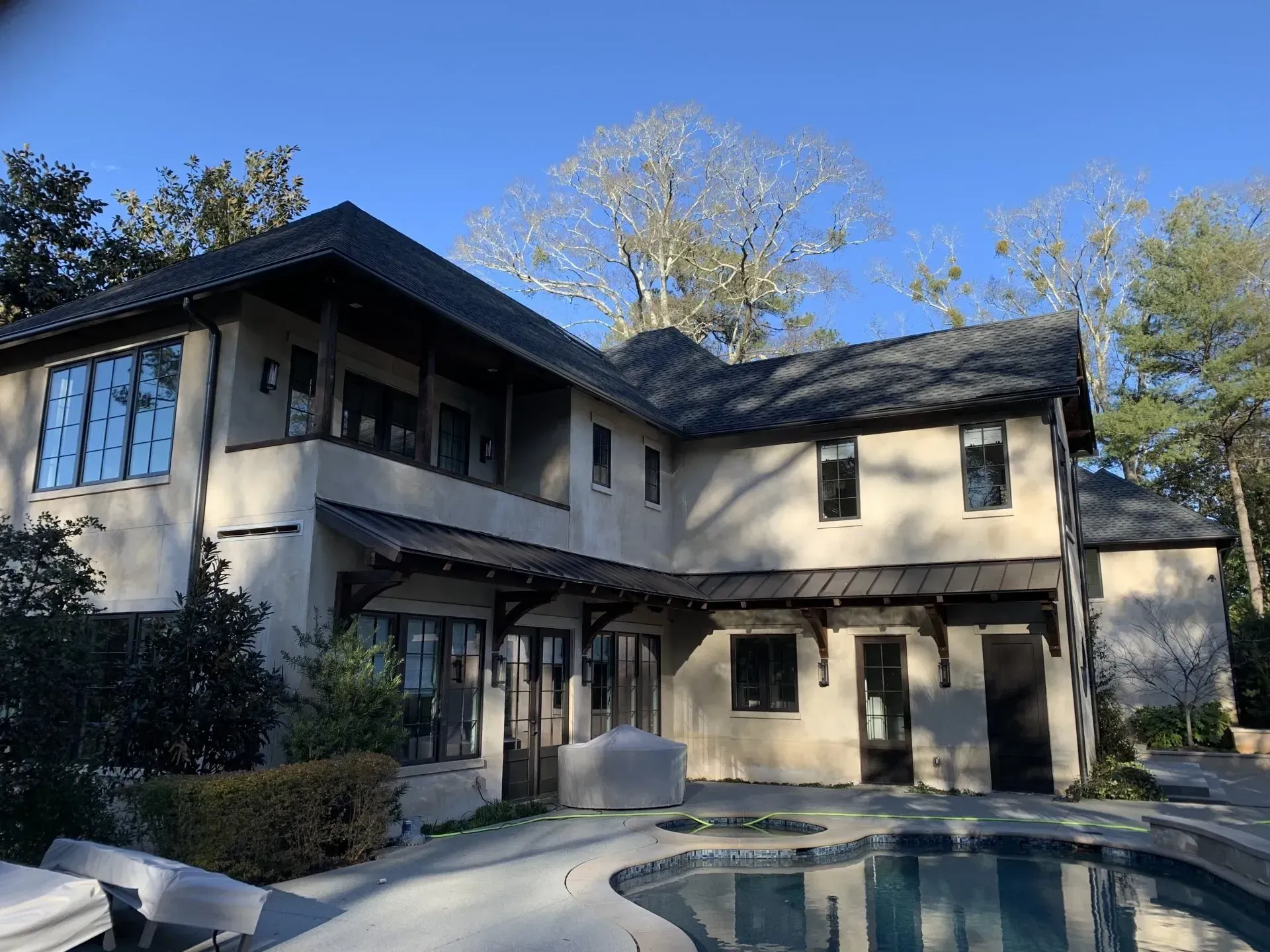 Two-story beige house with dark roof and trim, a pool in the foreground, and trees in the background.