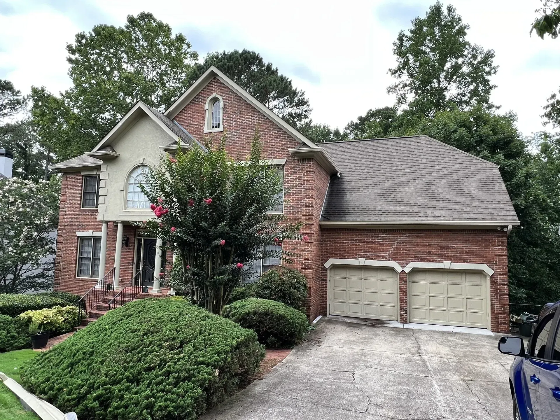 Brick house with a two-car garage, lush landscaping, and trees. Overcast sky.